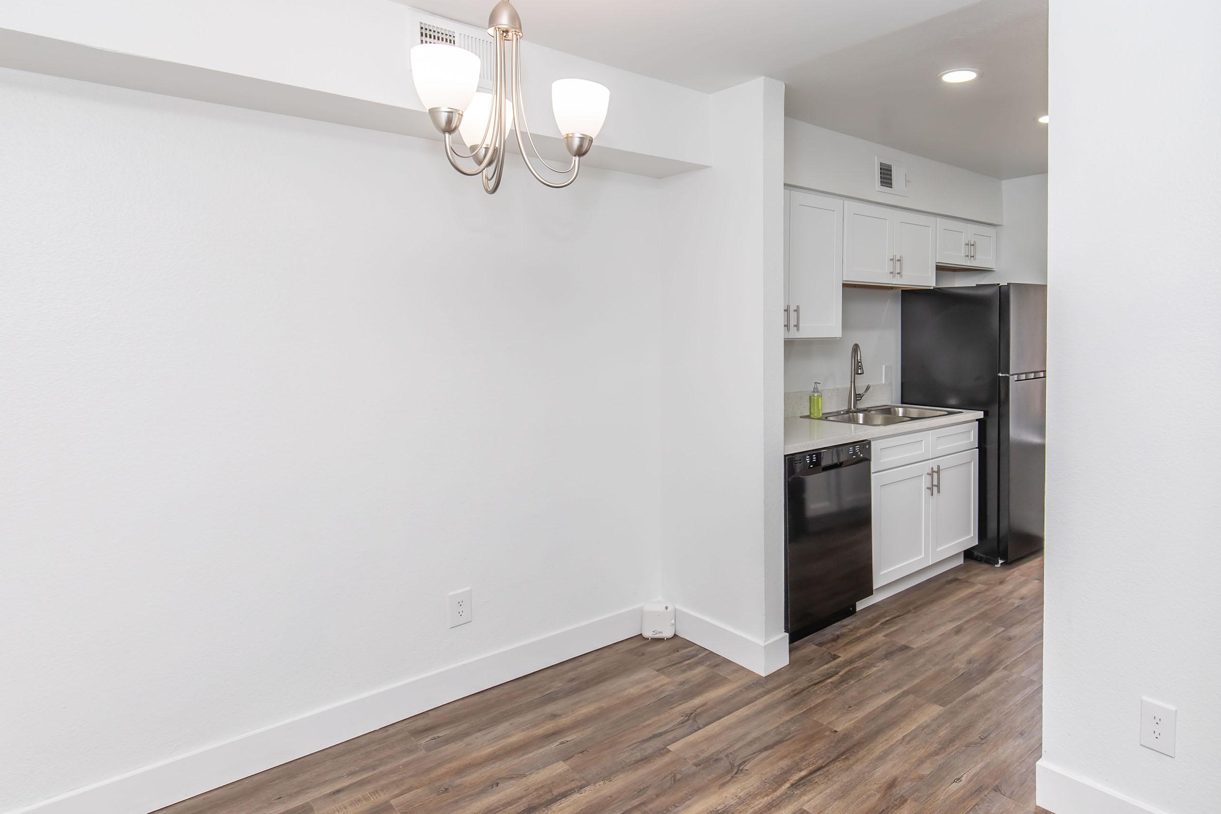 A modern kitchen area featuring white cabinetry, a stainless steel refrigerator, and a black dishwasher. There is a small dining area illuminated by a three-bulb chandelier. The floor is a wood-style laminate, and the walls are painted white, creating a bright and clean aesthetic.