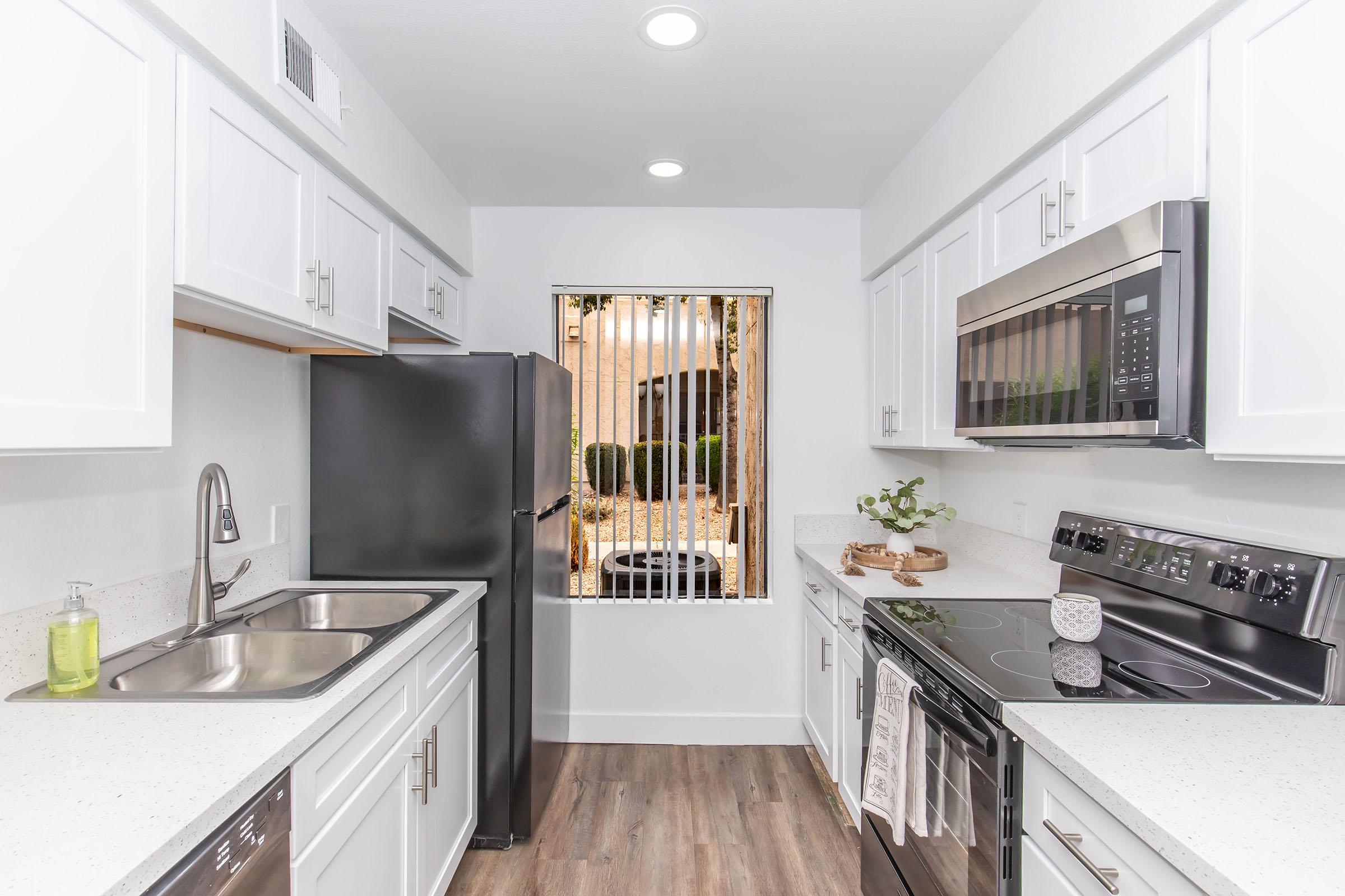 A modern kitchen featuring white cabinets, a black refrigerator, and stainless steel appliances including a microwave and oven. The countertops are light-colored with a sink on one side. A window with vertical blinds offers a view of outdoor greenery, adding natural light to the space.