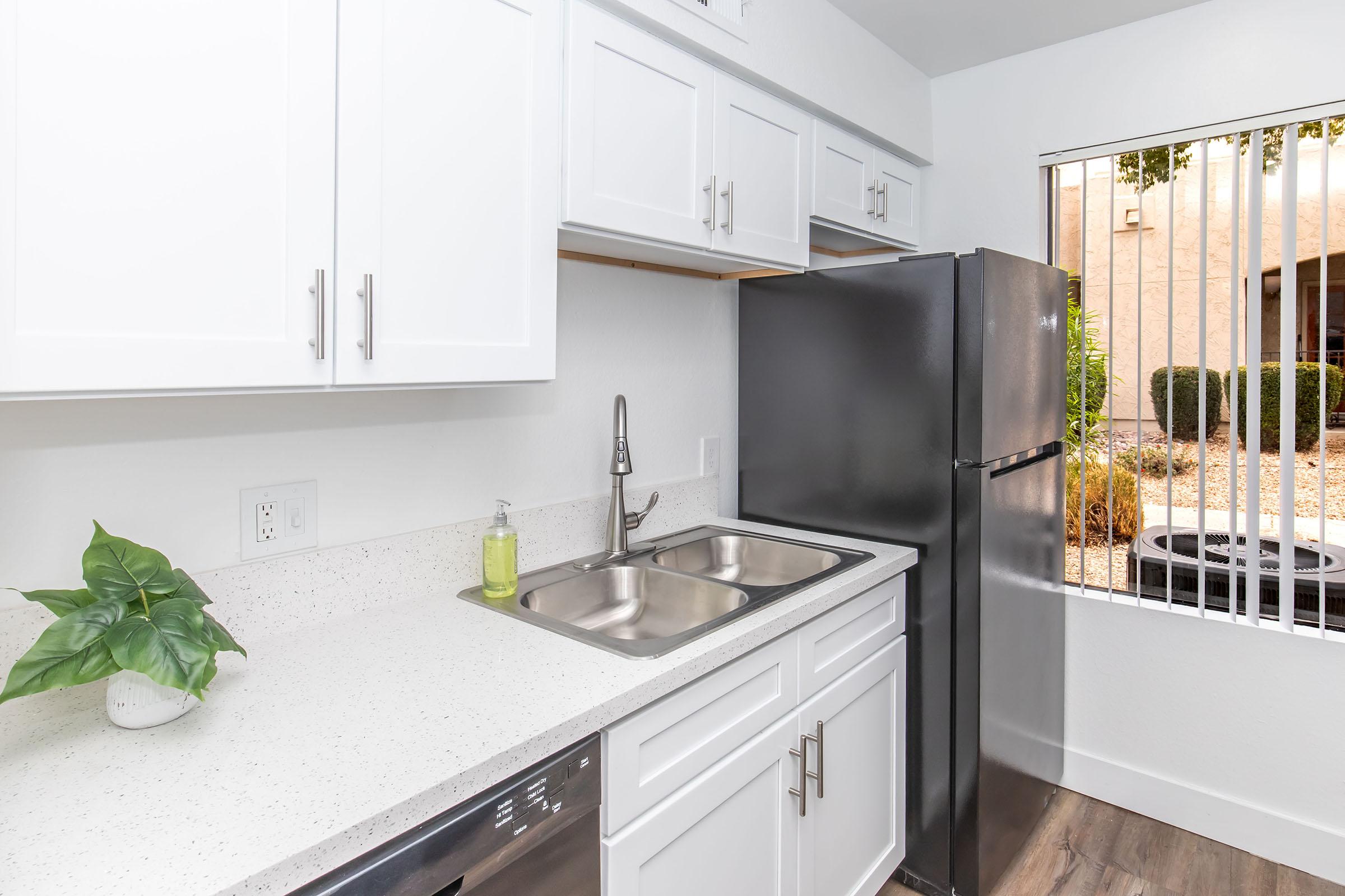 A modern kitchen with white cabinets, a double sink, and stainless steel appliances, including a refrigerator and a dishwasher. There is a small green plant on the countertop, and a window with blinds that offers a view of an outdoor area with shrubs and gravel.