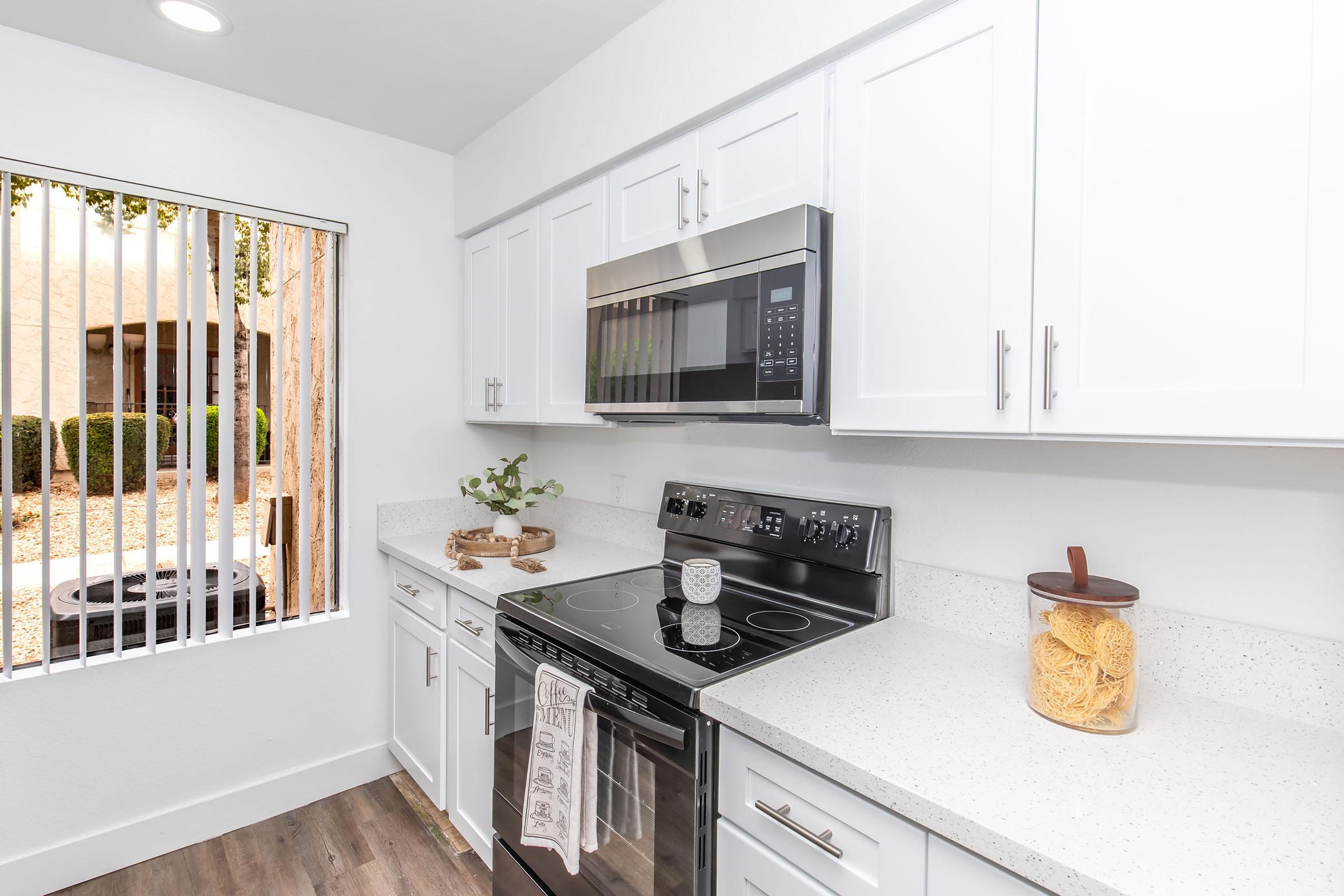 Modern kitchen featuring white cabinetry, a stainless steel microwave above an electric stove, and a quartz countertop. A window with vertical blinds provides natural light, showcasing outdoor greenery. Decorative elements include a plant and a jar of pasta on the counter.