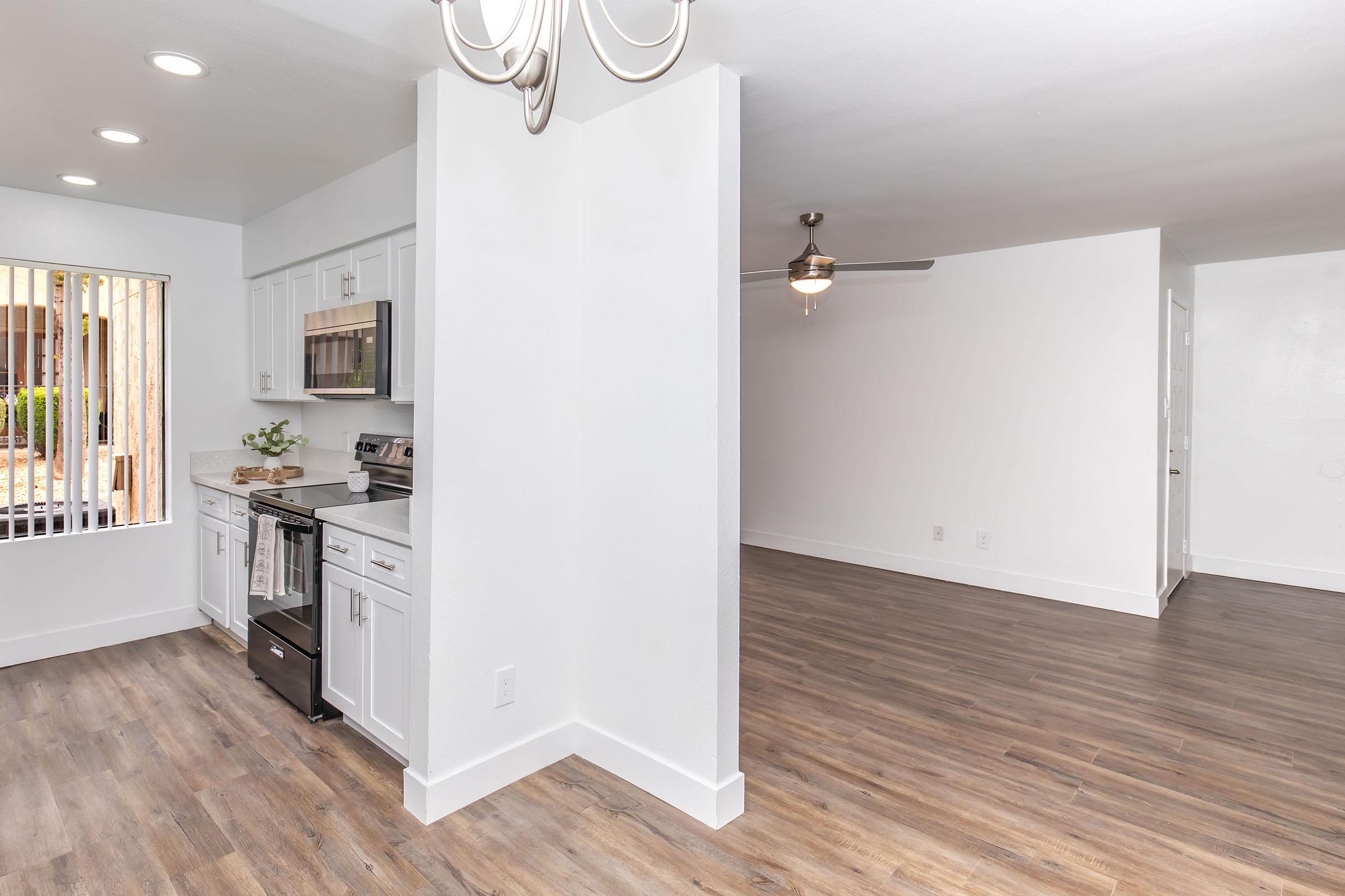 Modern kitchen and living area featuring white cabinetry, stainless steel appliances, and a ceiling fan. The space has a light, airy feel with large windows allowing natural light and a seamless flow to the adjoining room. The flooring is a warm wood laminate, enhancing the contemporary design.