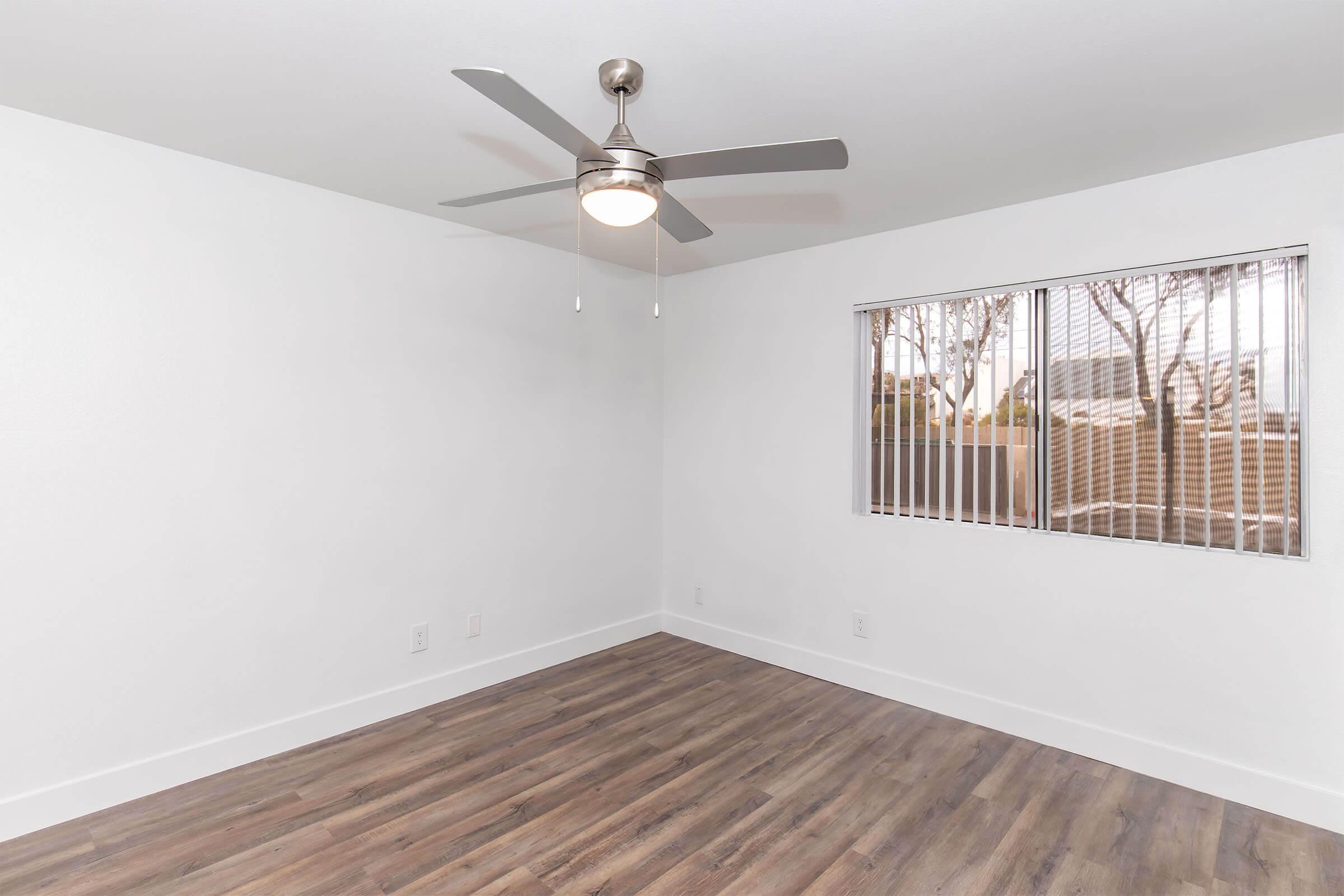 A bright, empty room with white walls and a ceiling fan. A window with vertical blinds lets in natural light, revealing a view of a fence outside. The floor is finished with light wood-like flooring, and there are no furnishings or decorations, creating a spacious and clean atmosphere.