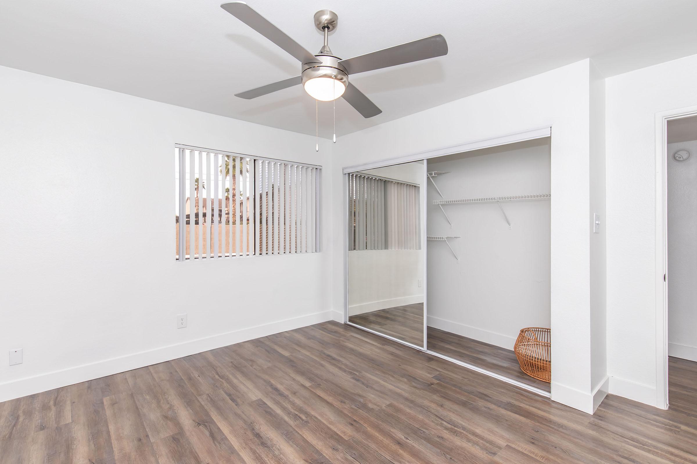 A spacious, bright bedroom featuring light-colored walls, a ceiling fan, and large sliding mirrored closet doors. The floor is covered with modern laminate flooring. Natural light streams in through a window with horizontal blinds, and a small wicker basket is positioned near the closet.