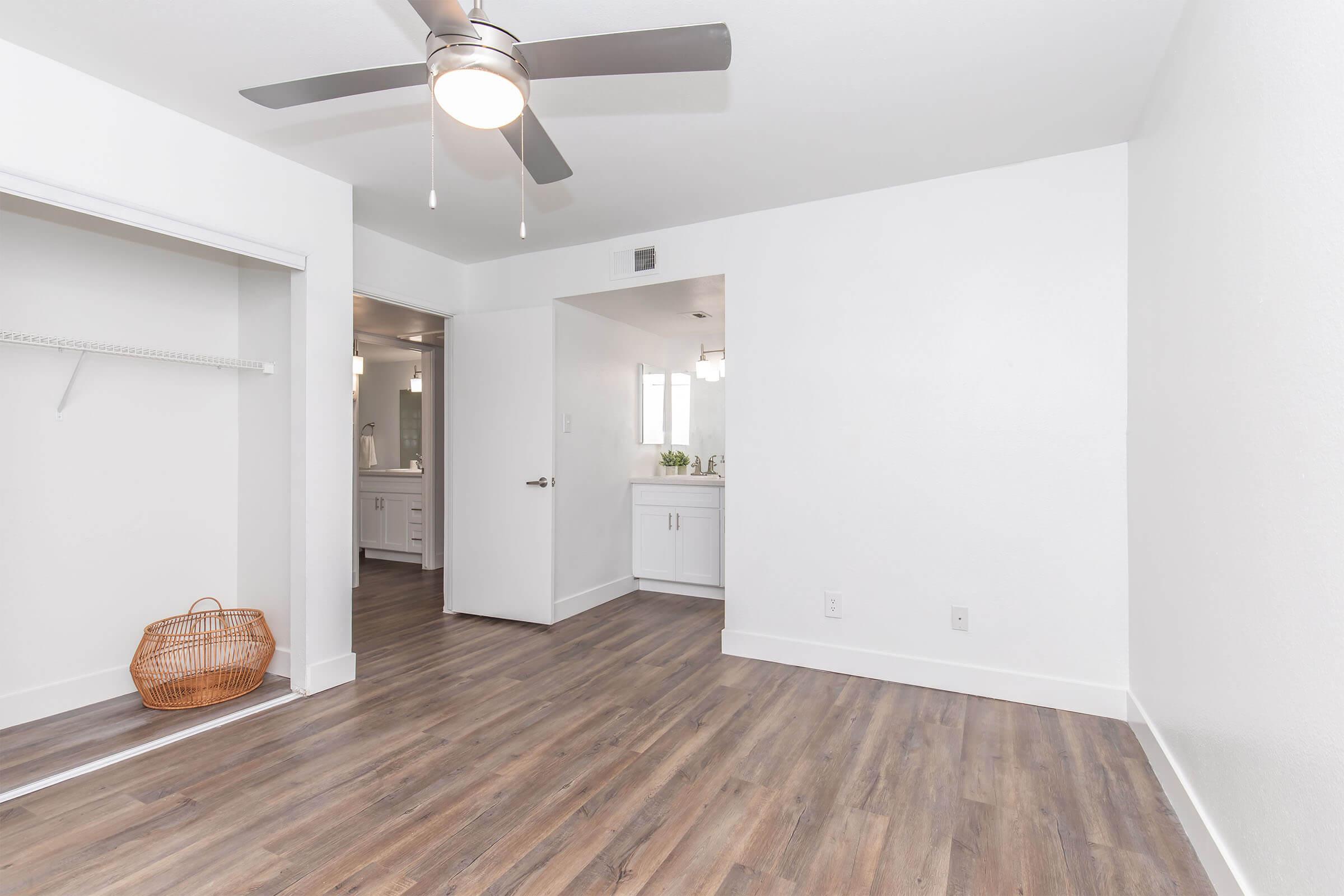 Bright, minimalistic room featuring light wood flooring, a ceiling fan, and a closet with a wicker basket. The space is open and airy, with a doorway leading to a bathroom and a neutral color palette, creating a modern and inviting atmosphere.