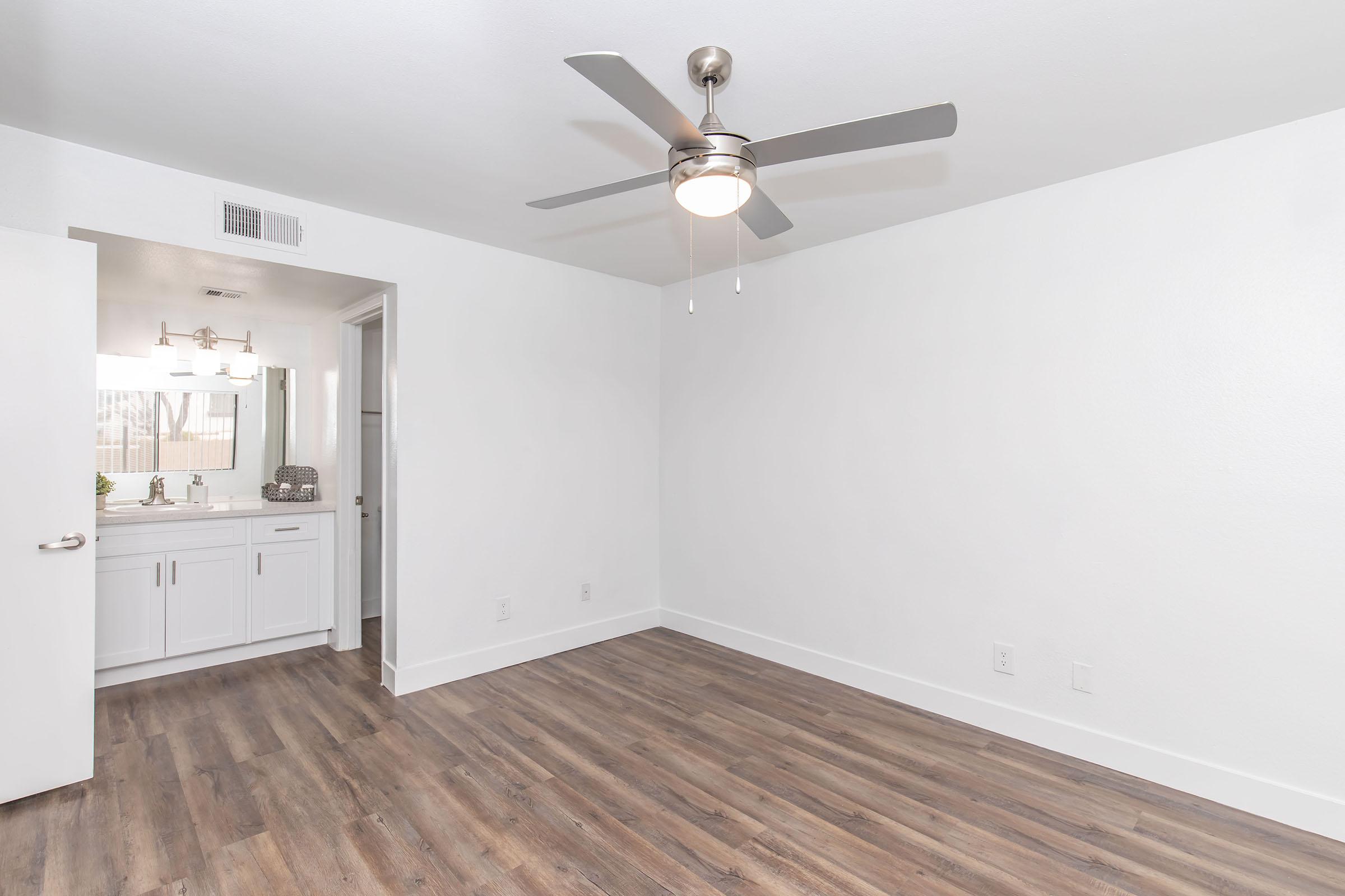A bright, modern room featuring a ceiling fan, beige hardwood flooring, and a door leading to a bathroom with white cabinetry. The walls are painted white, creating a clean and spacious feel. No furniture is present, emphasizing the room's emptiness. Natural light flows in, enhancing the inviting atmosphere.