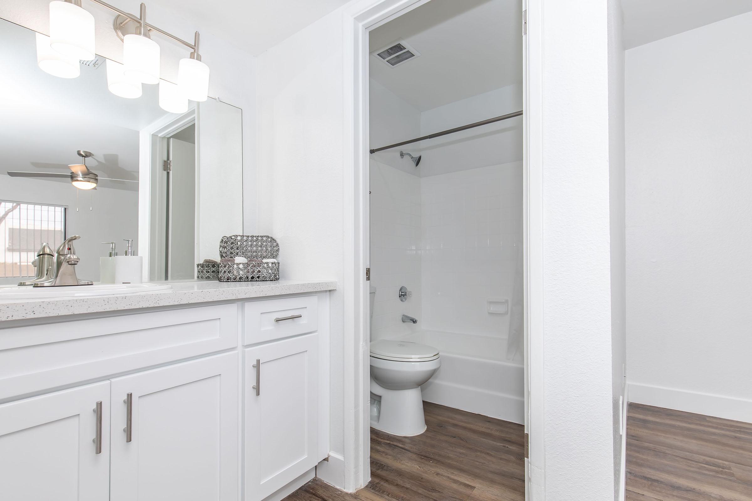 A well-lit bathroom featuring a modern sink with a mirror, white cabinetry, and a shower-tub combo. The walls and fixtures are predominantly white, and the floor has a wood-like appearance. A glimpse of a ceiling fan is visible, indicating a spacious layout.