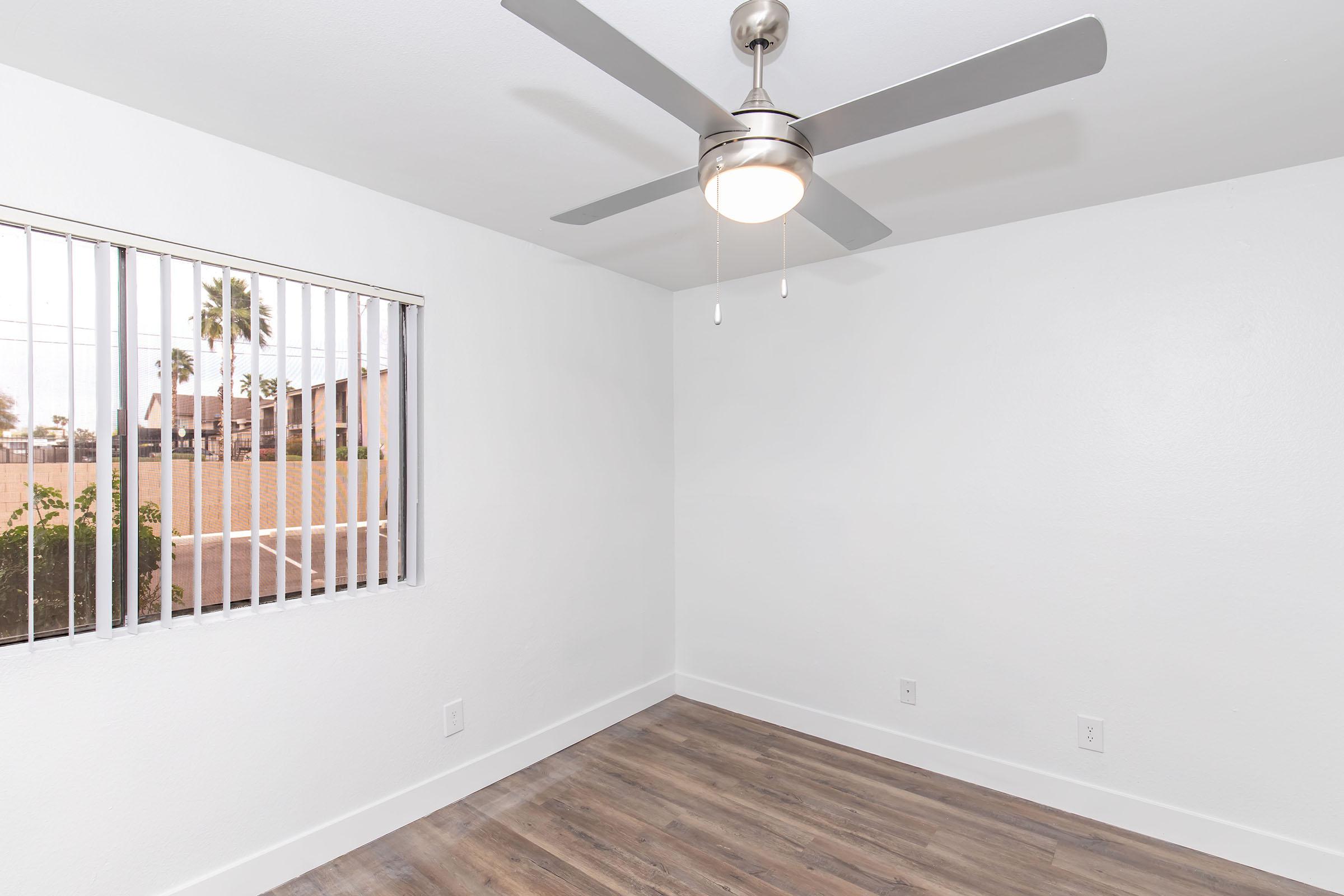 A bright, empty room featuring light grey walls and wooden flooring. A ceiling fan with a light fixture is installed in the center. A window with vertical blinds allows natural light in, showing a view of a garden outside. The overall ambiance is clean and modern.