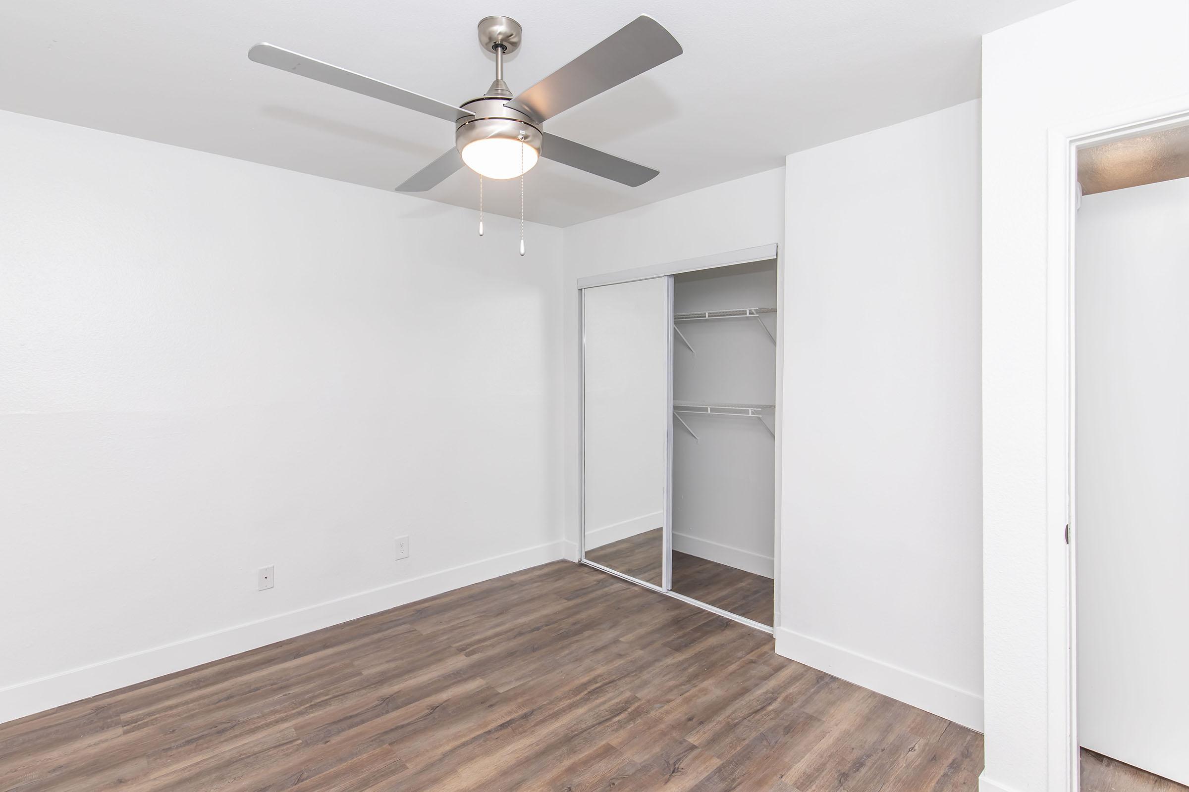 A spacious, well-lit room with light-colored walls and a ceiling fan. The floor features wooden laminate, and there is a mirrored closet with sliding doors. A door on the right leads to another area, enhancing the room's open feel. Natural light brightens the space, creating a welcoming atmosphere.