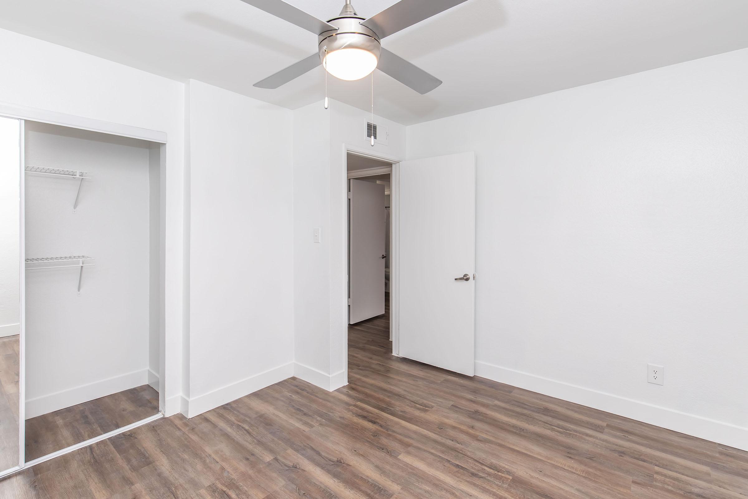 A well-lit, minimalistic room featuring light-colored walls, a ceiling fan, and laminate flooring. A mirrored closet is on the left side, while the open door leads to another room in the background. The overall ambiance is clean and modern.