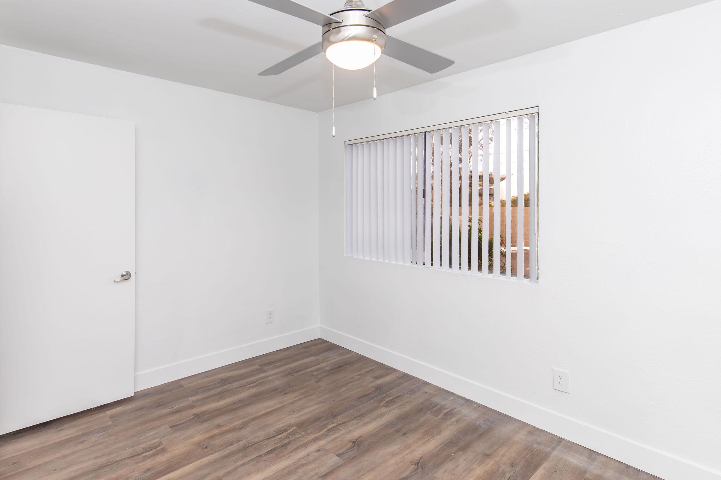 A bright, minimalist room featuring a ceiling fan, light-colored walls, and a window with vertical blinds. The floor is made of wood-like laminate, and there is a closed white door on the left. The space is clean and uncluttered, creating a fresh and airy atmosphere.