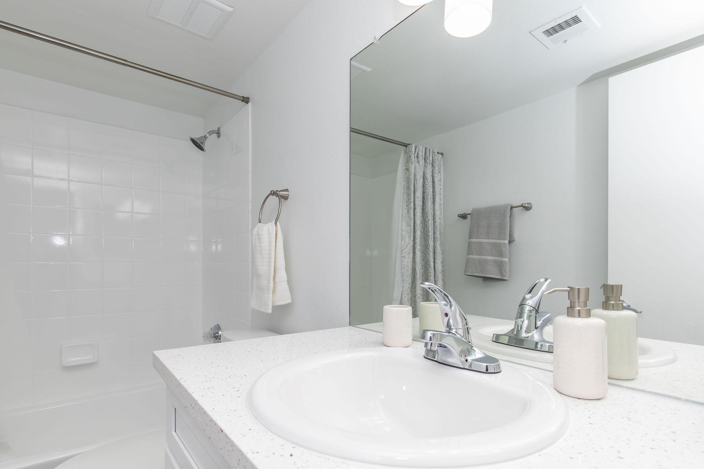 A clean and modern bathroom featuring a white countertop with a sink, silver faucets, and two soap dispensers. In the background, there is a shower area with a glass door, a towel hanging on a rack, and a large mirror reflecting the space. The walls are painted in a light color, contributing to a fresh atmosphere.