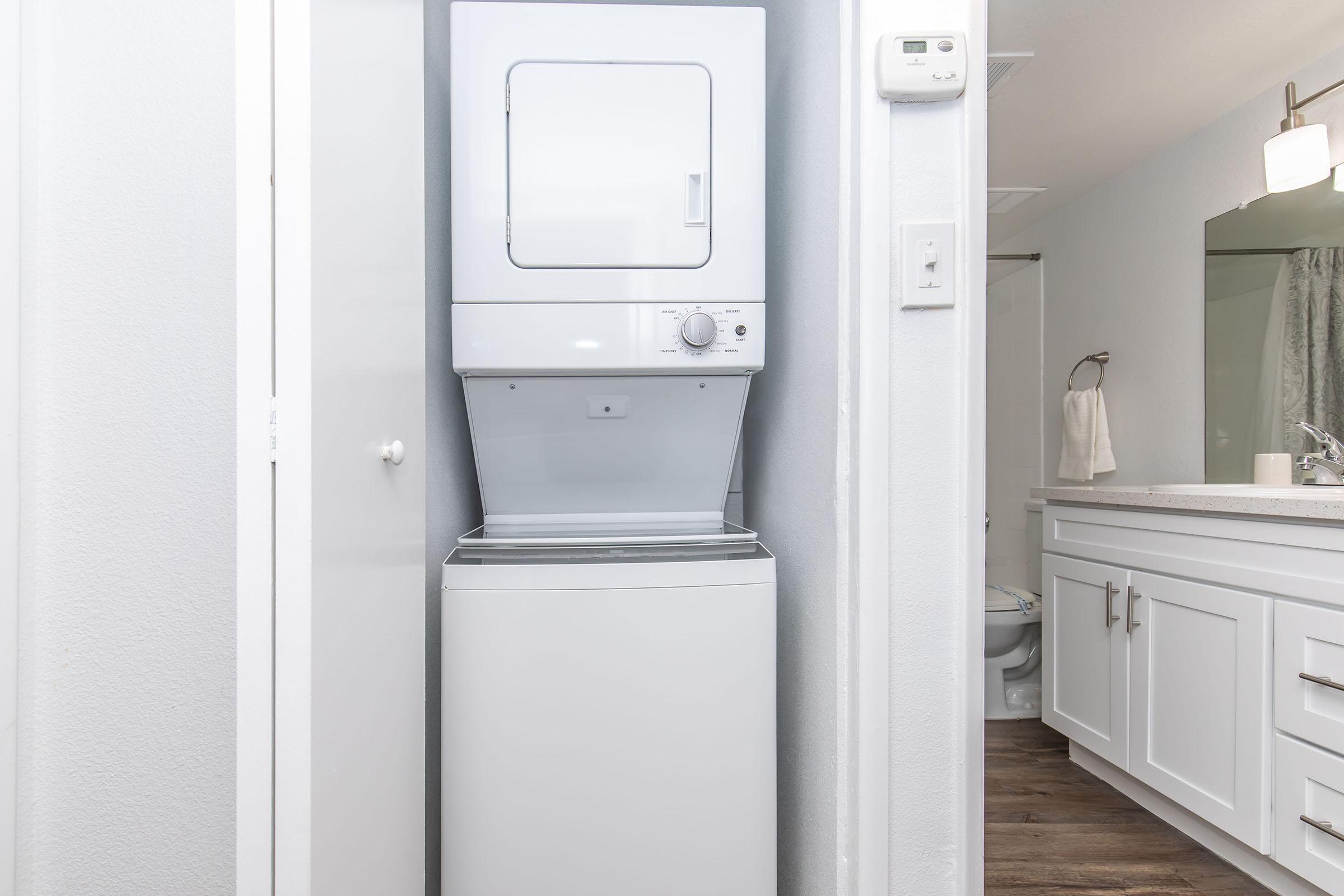 A compact laundry area featuring a stacked washer and dryer unit against a light gray wall, with a white cabinet door next to it. In the background, a bathroom is visible with a sink and mirror, along with a towel hanging on a hook. The overall setting appears clean and modern.