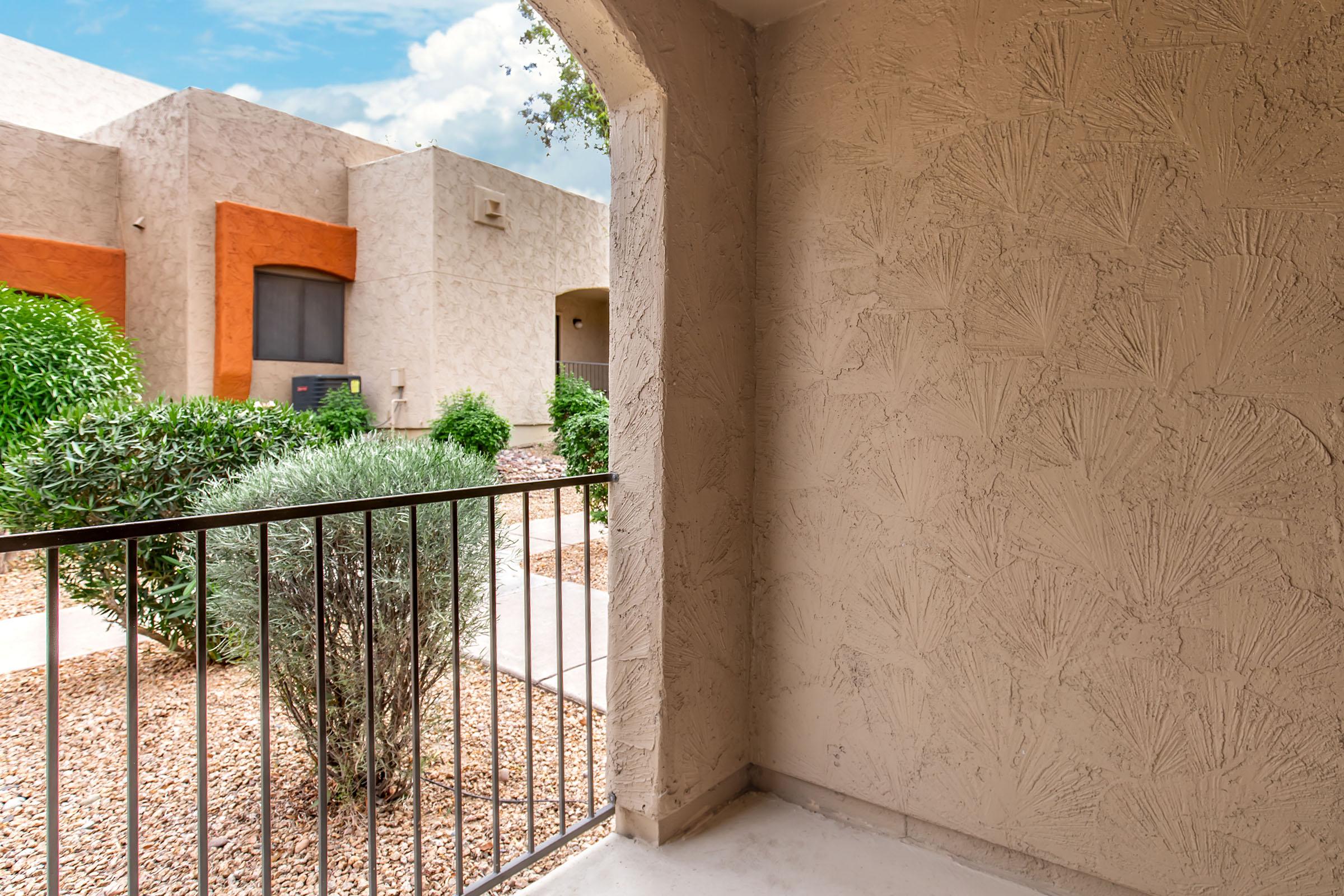 View from a balcony showcasing a landscaped courtyard with shrubs and small trees. The wall features a textured surface, and glimpses of neighboring buildings with varied colors are visible in the background under a partly cloudy sky.