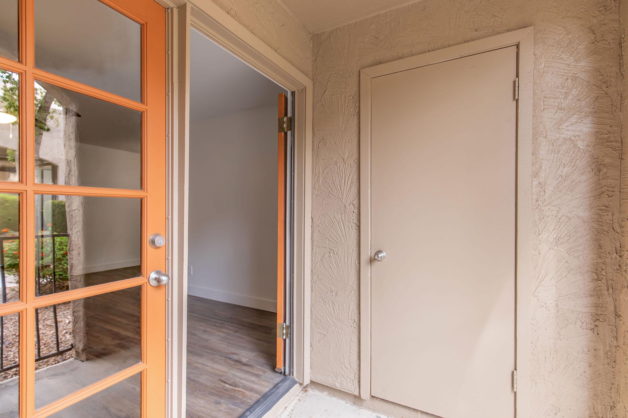 A view of a door entrance with orange-framed glass doors leading into a room. Adjacent is a closed beige door. The entryway features textured walls and visible flooring. Natural light comes through the glass doors, creating a welcoming atmosphere.