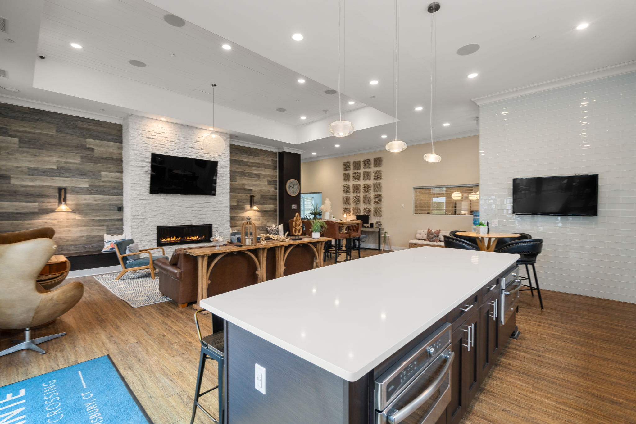 A modern living space featuring an open kitchen with a white countertop, stylish pendant lights, and a wooden accent wall. The room includes a cozy seating area with leather couches, a fireplace, and large windows. There is a television mounted on the wall, and decorative elements add a warm ambiance.