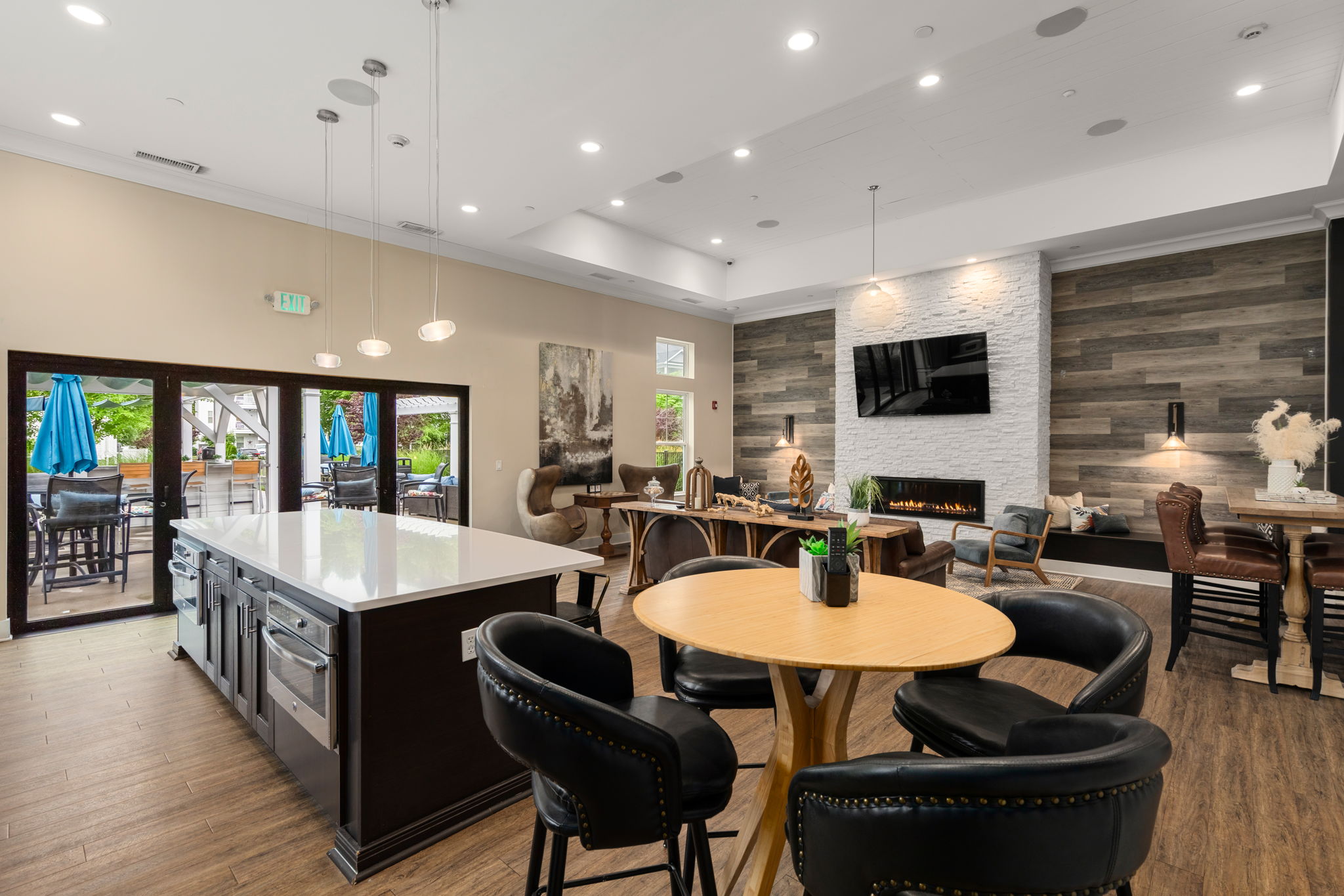 A modern communal space featuring a large kitchen island with bar stools, a dining table, and a cozy seating area. Large windows provide natural light, and there are various decorative elements, including artwork and plants. The space opens to an outdoor area with blue umbrellas.