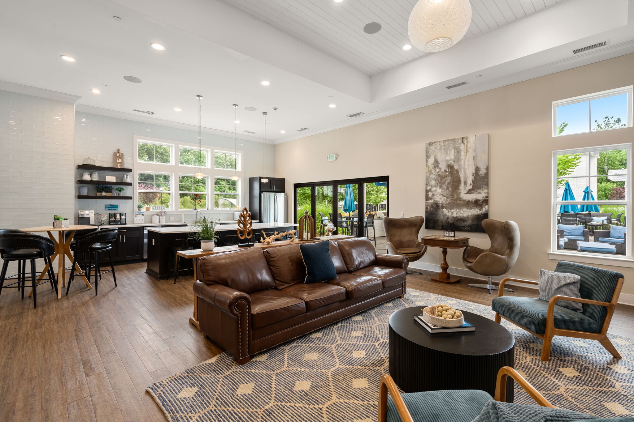 Modern lounge area featuring a brown leather sofa, wooden coffee table, and stylish armchairs. The space has large windows allowing natural light, a kitchen area with bar stools, and contemporary artwork on the walls. The decor includes a geometric rug and a light fixture, creating a welcoming atmosphere.