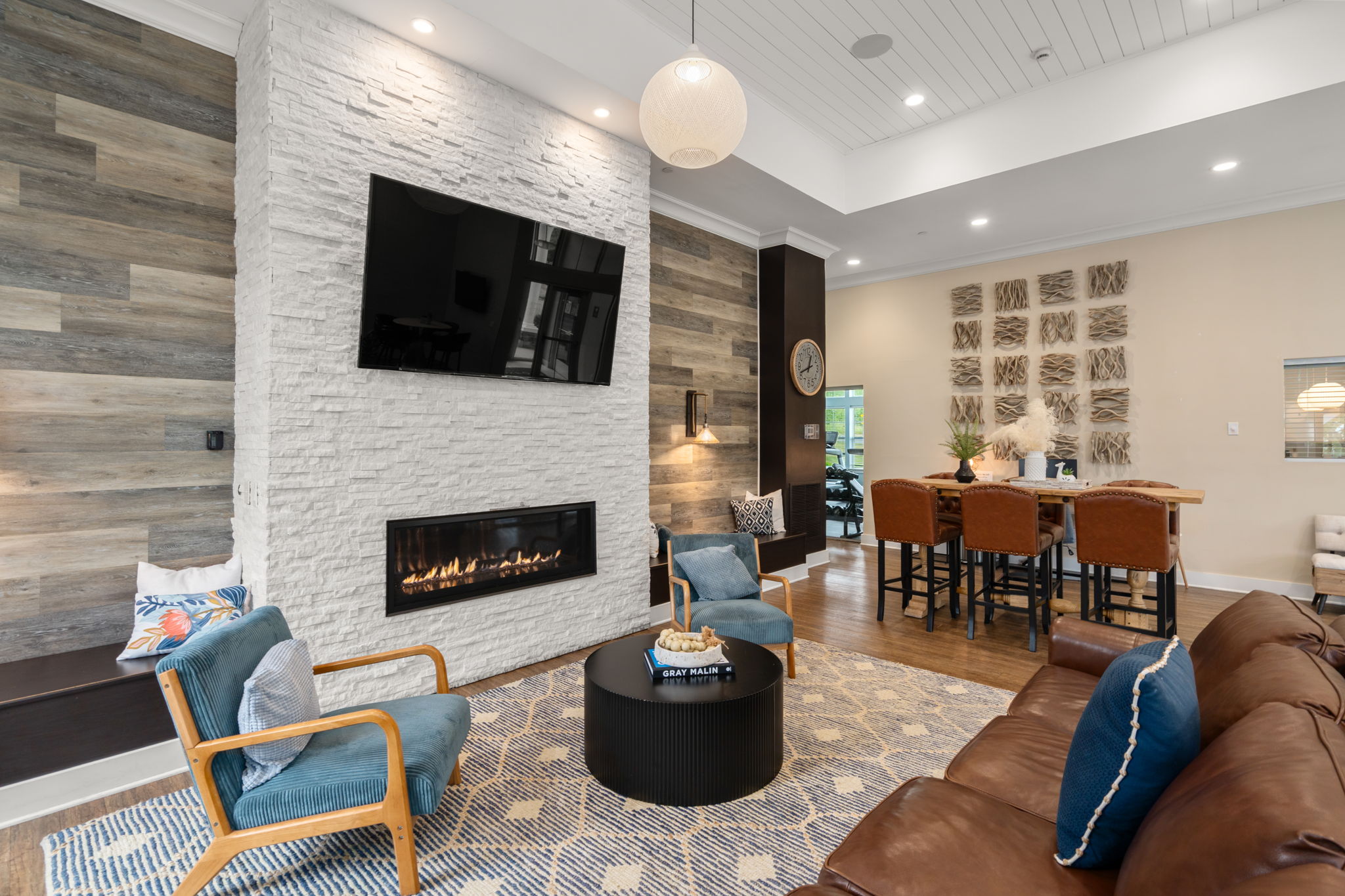 A modern living room featuring a cozy seating area with a brown leather couch and blue accent chairs. A white stone fireplace with a mounted TV is the focal point, surrounded by wood-paneled walls. In the background, there's a dining area with a table and chairs, creating an inviting and stylish atmosphere.