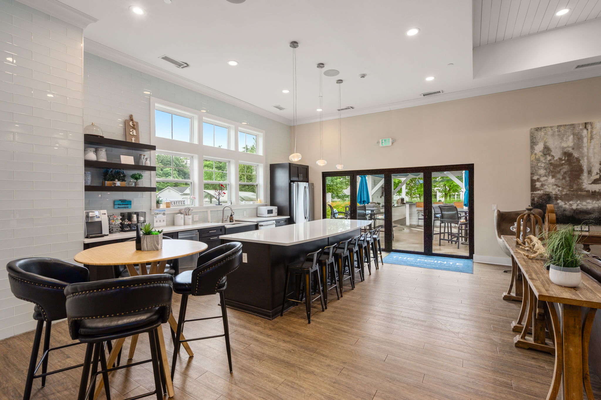 A modern, spacious lounge area featuring a large kitchen island with barstools, wooden flooring, and large windows allowing natural light. On one side, there are shelves with decor and appliances, while the other opens to an outdoor patio with seating. The overall design is contemporary and inviting.