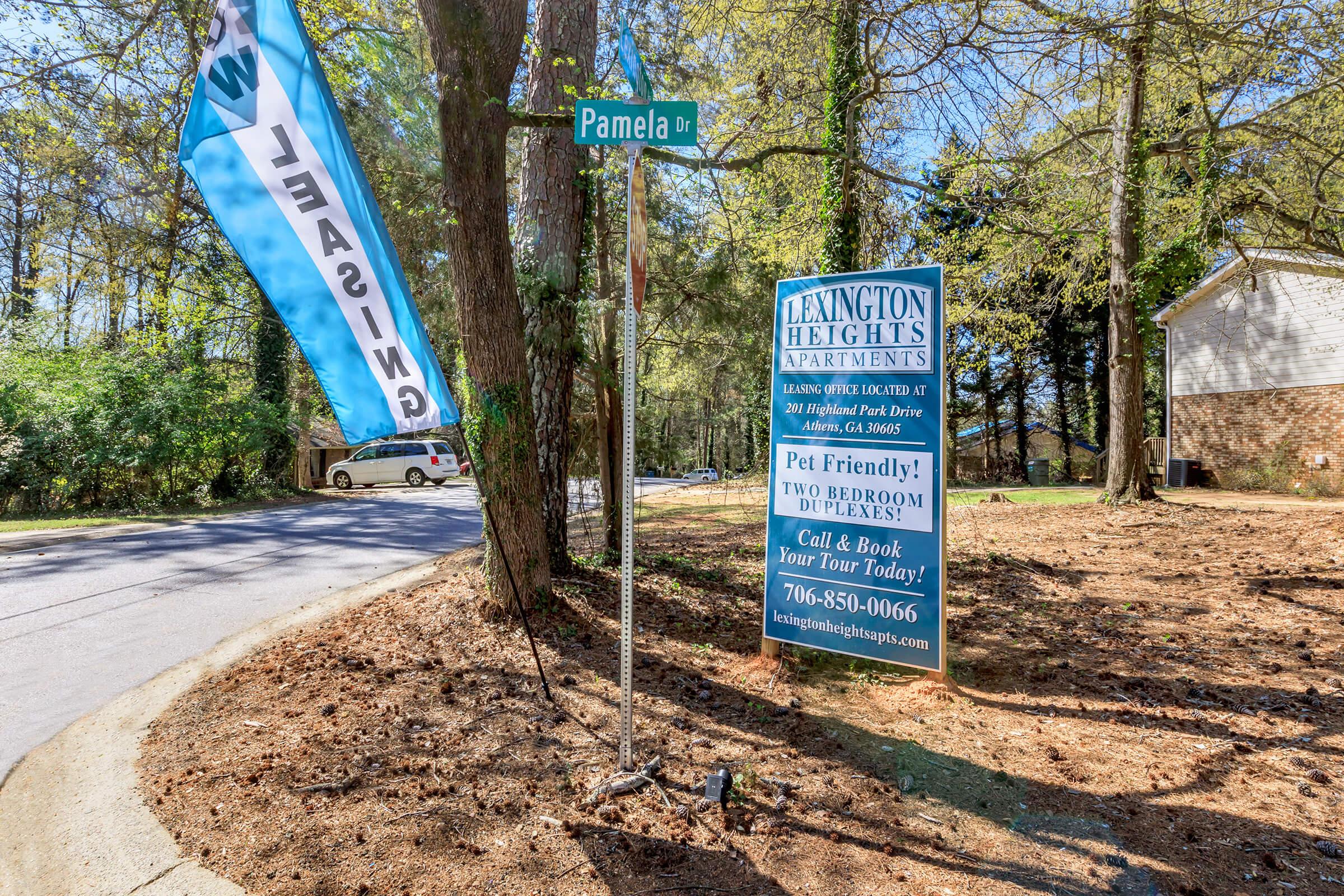 A street sign for Pamela Drive at the intersection with a banner flag indicating "Leasing." Nearby, a large sign advertises Lexington Heights Apartments, highlighting features such as pet-friendliness and two-bedroom units, along with a contact number for inquiries and bookings.