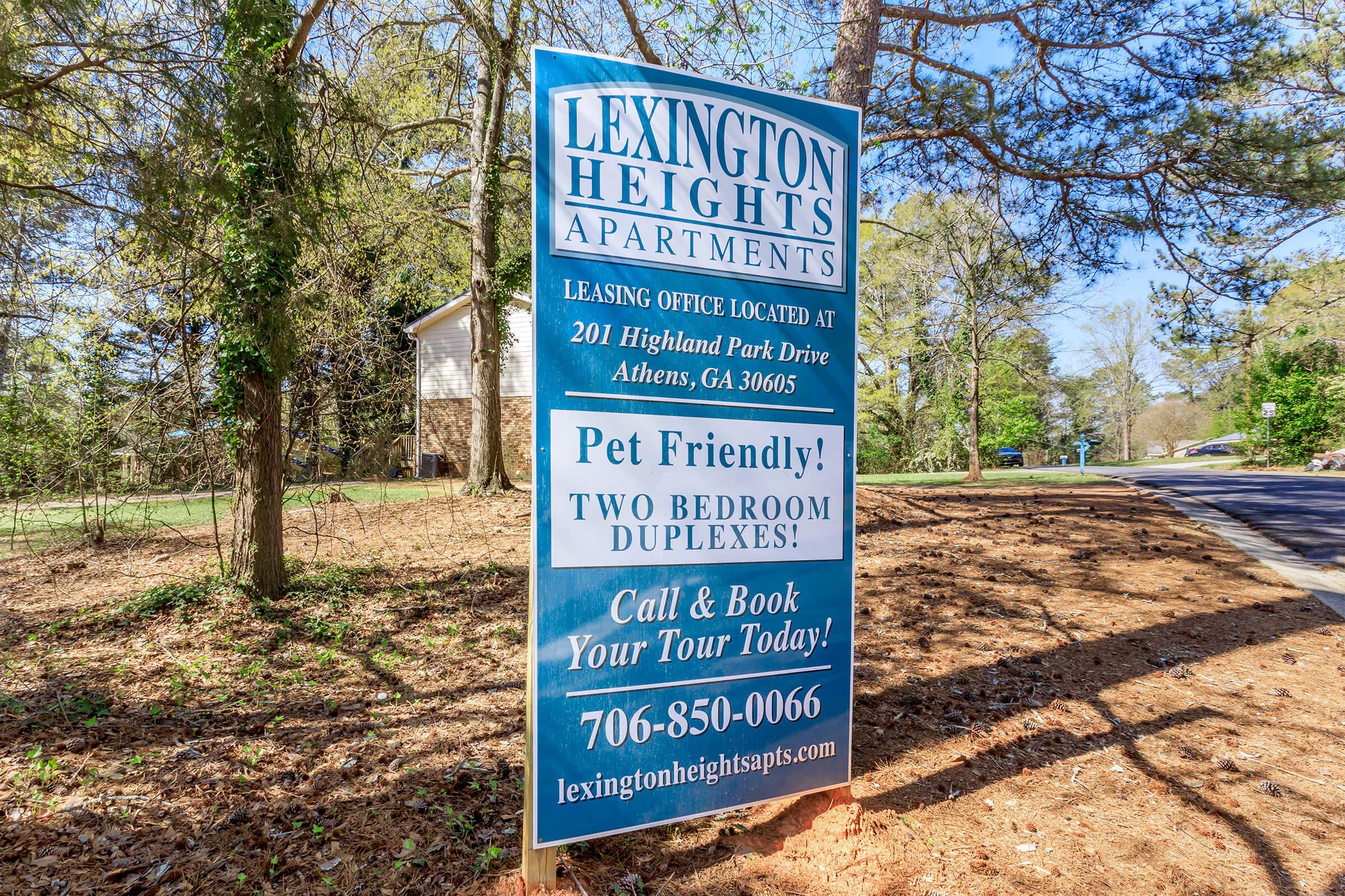 Sign for Lexington Heights Apartments, featuring information about the leasing office's location, contact details, and a promotion for pet-friendly two-bedroom duplexes. Includes a call to action to book a tour. The setting is surrounded by trees in a residential area.