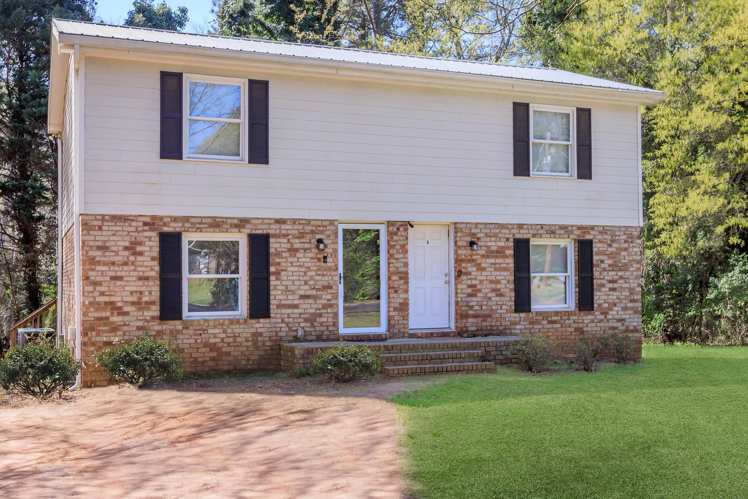 Two-story house with white siding and brick accents. The front features a white door, two windows on the upper level, and two on the lower level, all framed with black shutters. Neatly landscaped yard with green grass and small bushes, surrounded by trees in the background.