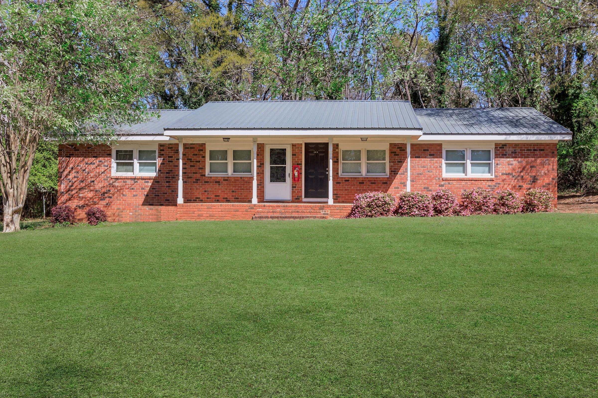 A charming single-story brick house with a gray metal roof and white-framed windows, set on a well-maintained green lawn. Flowers and shrubs line the front, and trees provide a backdrop. The house features a welcoming entrance with two doors and symmetrical window placement.
