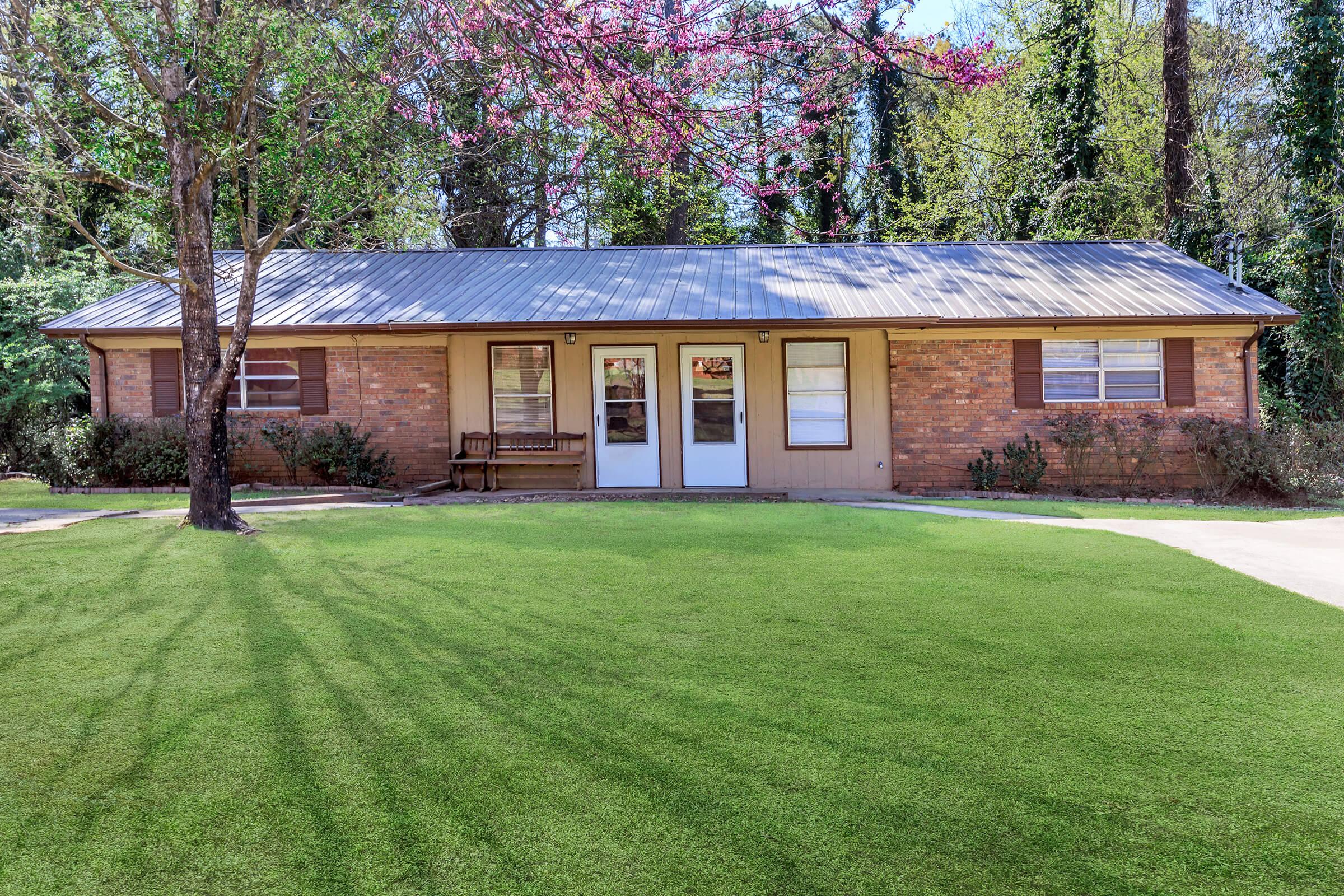 A single-story brick house with a metal roof, featuring two front doors and several windows. A well-maintained lawn surrounds the house, with a tree and shrubs in the front yard. Bright green grass and blooming trees in the background add to the serene atmosphere.