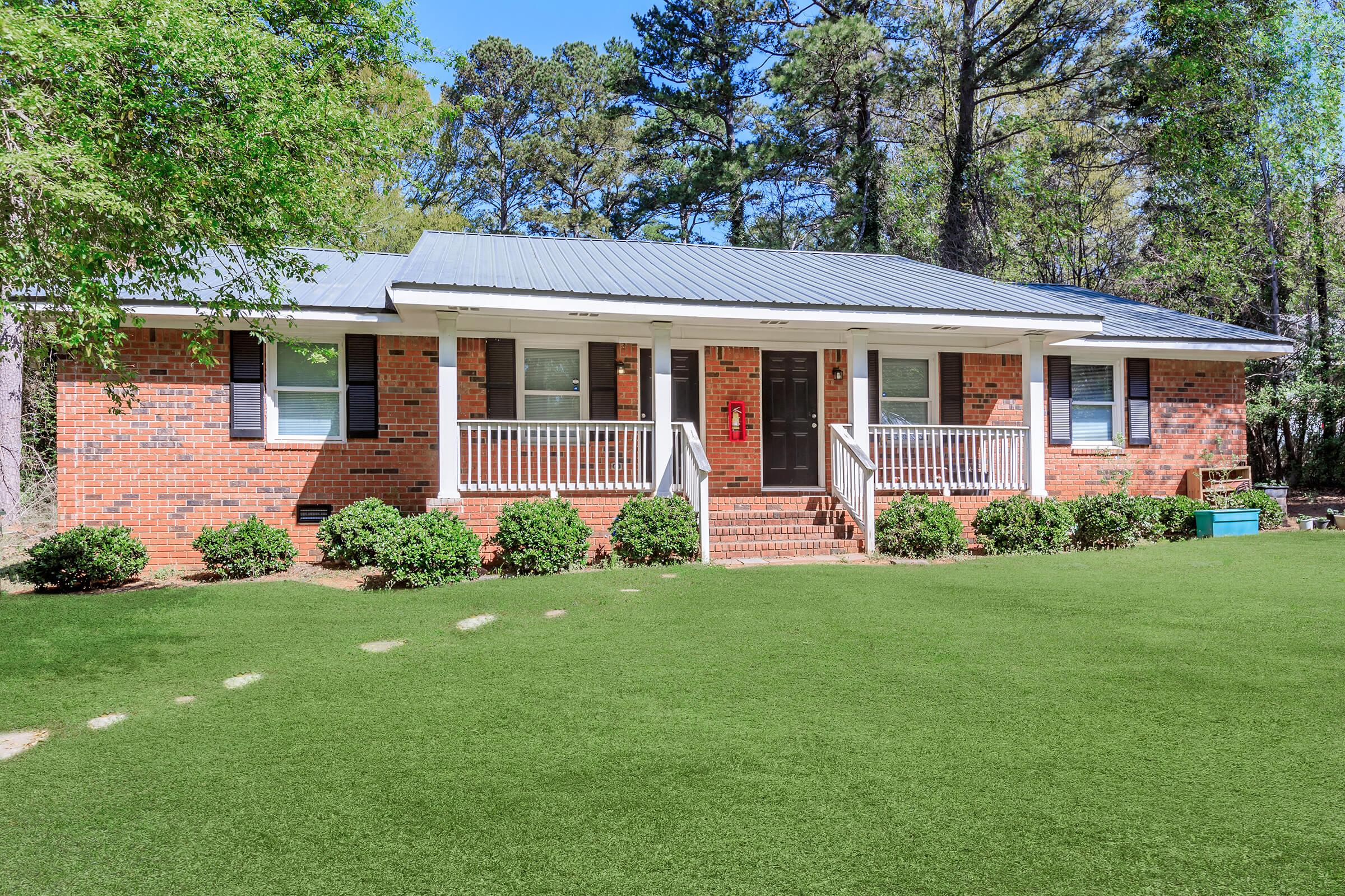 Single-story brick house with a gray metal roof, white railing on the porch, and green lawn. Surrounded by trees, the front yard features small shrubs and a stone pathway leading to the entrance.