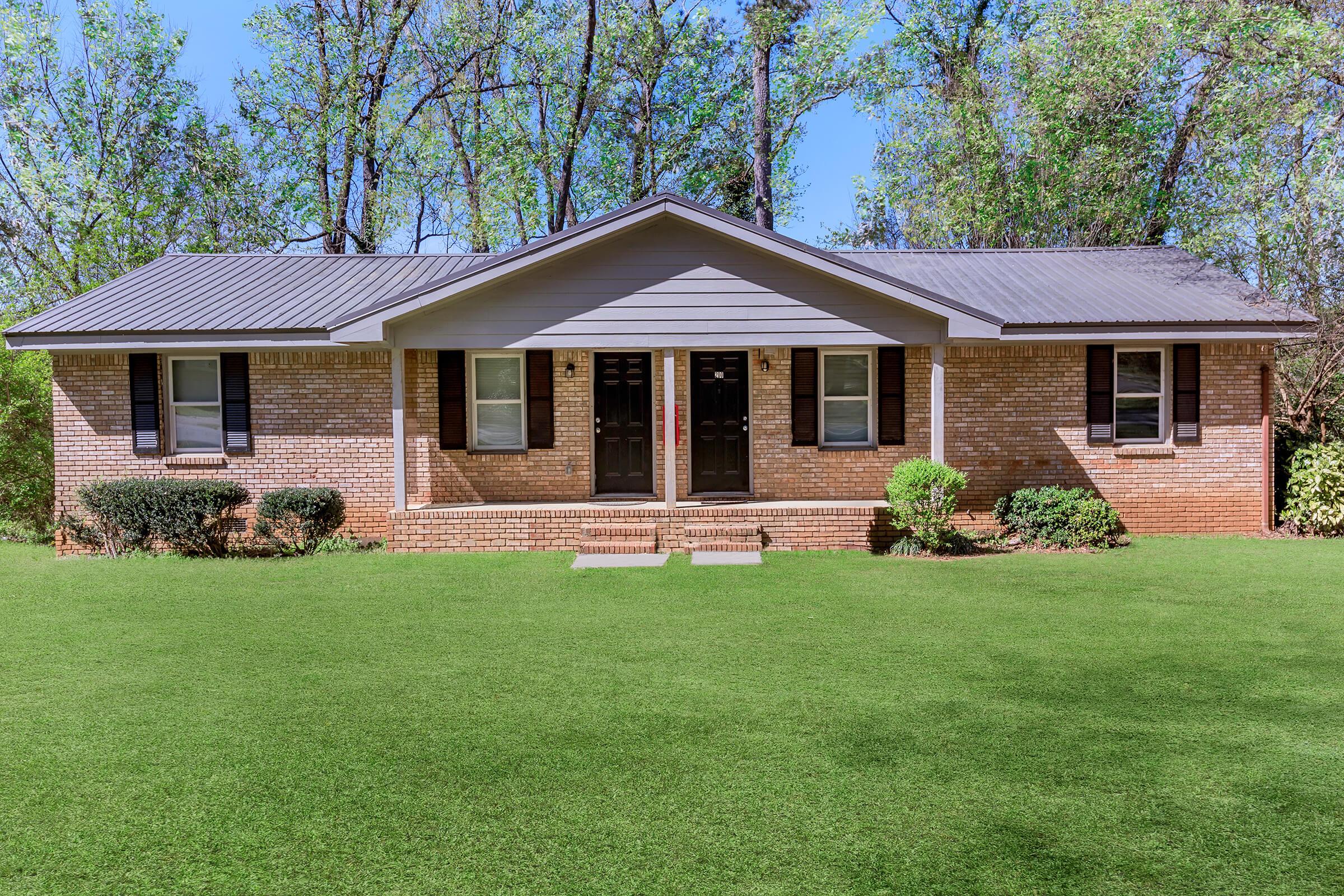 A single-story brick house with a gray roof, featuring two front doors and landscaped green grass. Surrounding the house are bushes and trees, indicating a lush outdoor setting in a residential area. The scene is bright and well-lit, showcasing a clear blue sky overhead.