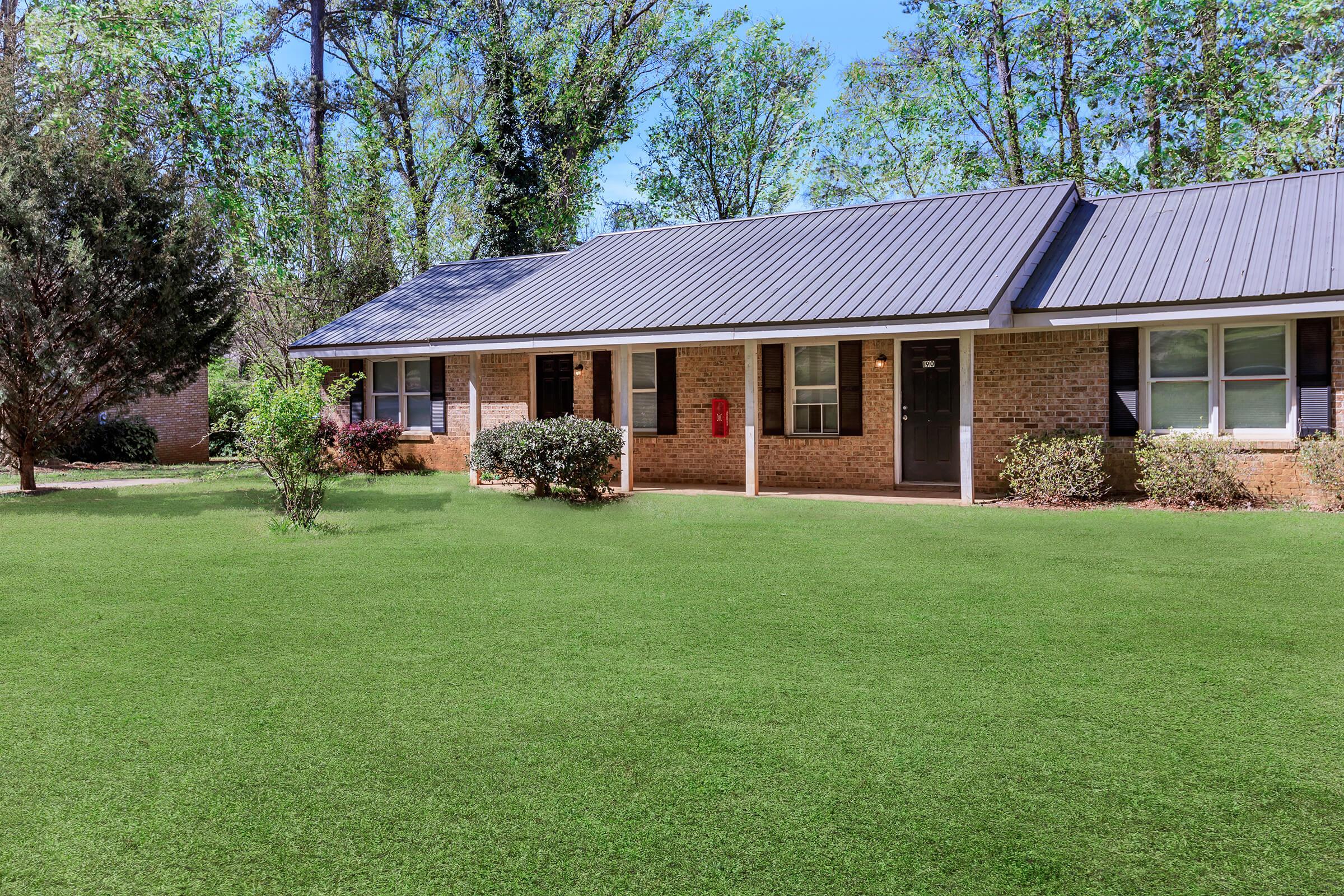 A well-maintained brick apartment building with a gray metal roof, featuring multiple entrances. The building is surrounded by a lush green lawn and small shrubs. Trees can be seen in the background, indicating a serene, landscaped environment.