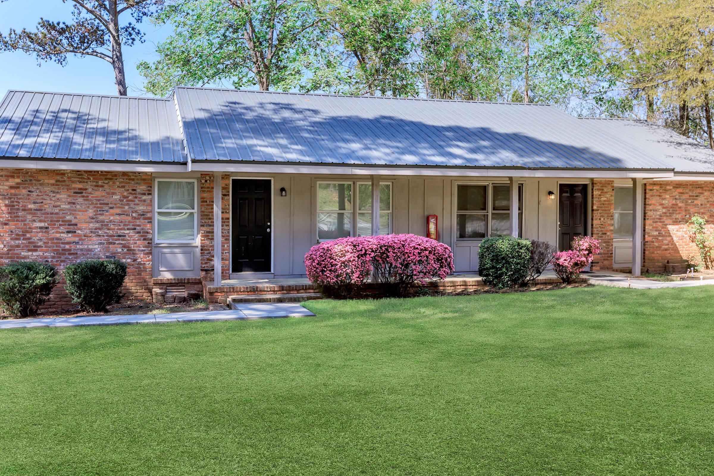 Two side-by-side residential units with brick exteriors and a gray metal roof. Each unit has a front door, one black and one white, bordered by neatly trimmed bushes and vibrant pink flowering plants. A well-maintained green lawn stretches in front of the homes, with trees and foliage in the background.