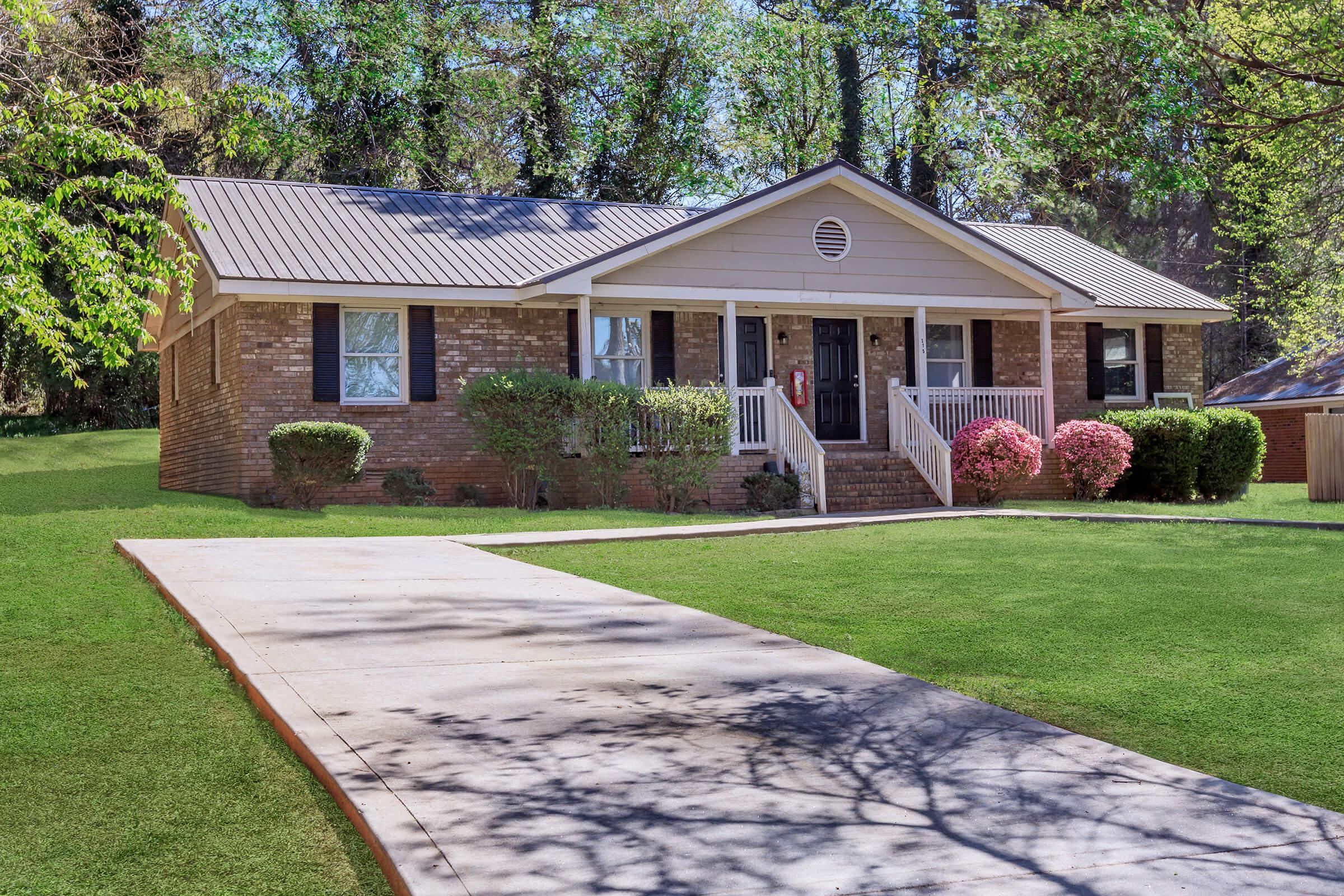 A single-story brick house with a gray metal roof, featuring a front porch with white railings, surrounded by green grass and neatly trimmed bushes. Colorful flowering plants are visible near the entrance, and the paved driveway leads up to the house. Sunlight creates shadows on the lawn.