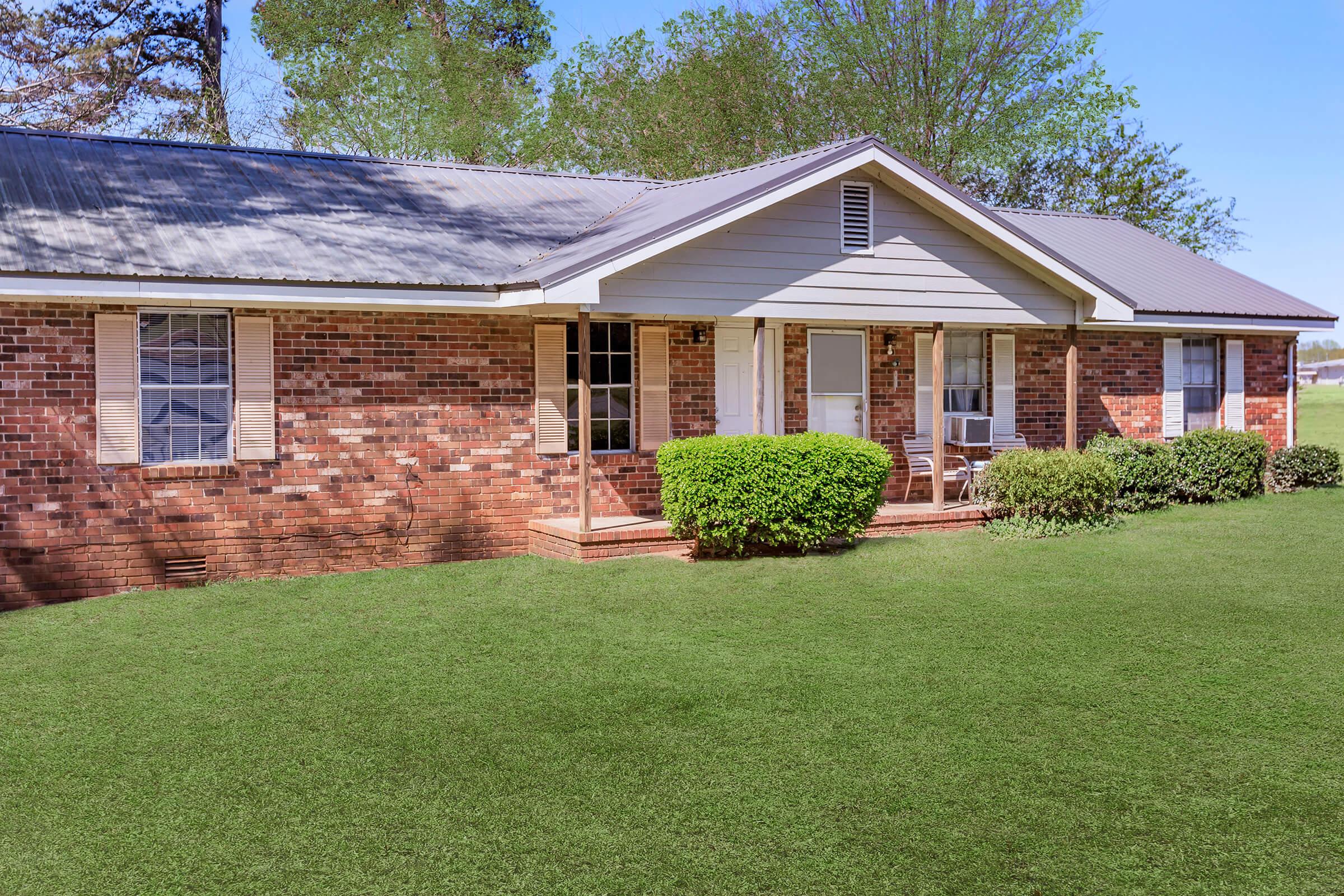 A single-story brick house with a peaked roof, featuring white shutters and a small porch. The front yard is neatly maintained with green grass and shrubs. Trees line the background, and the sky is clear and blue.