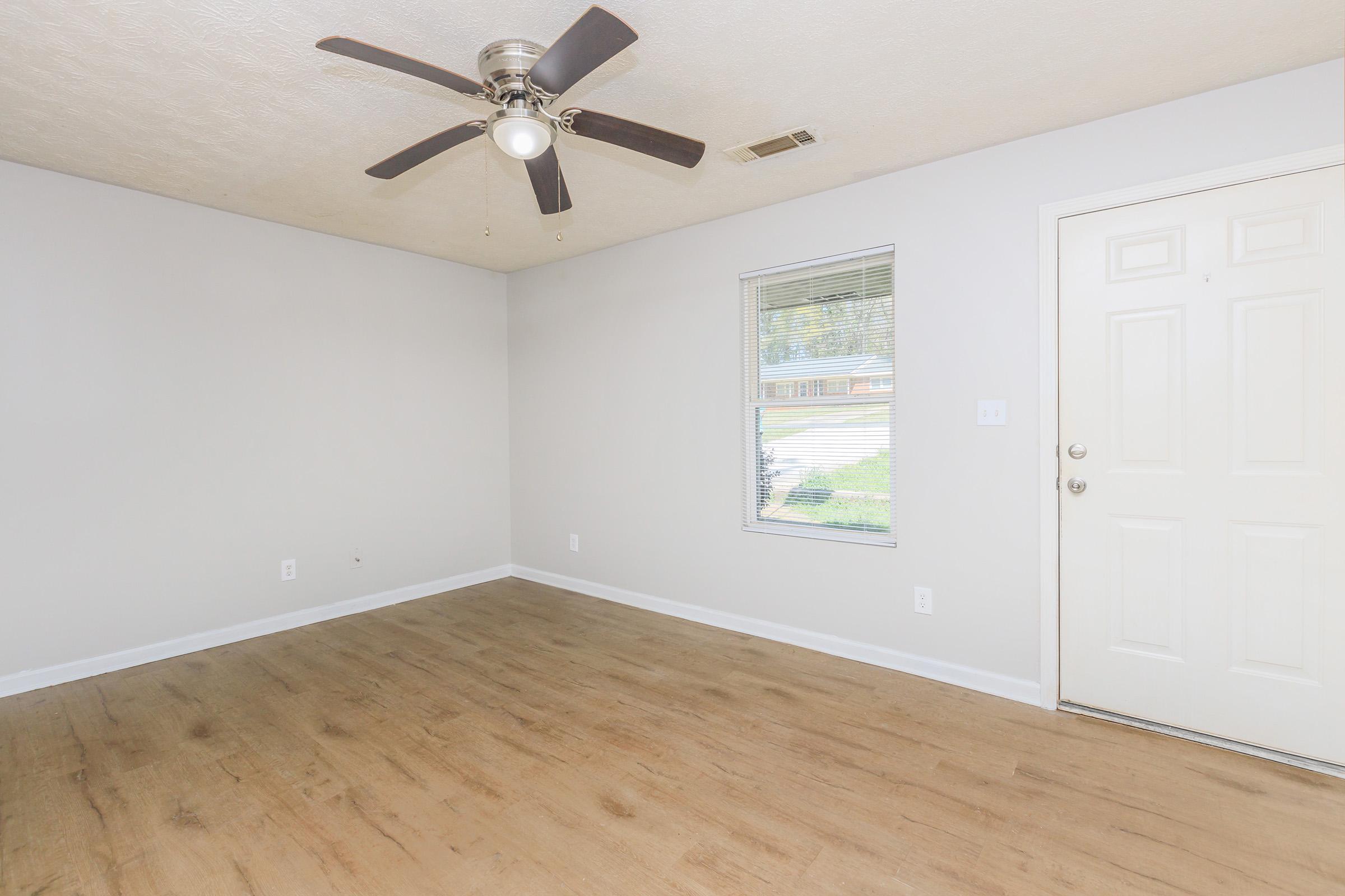 Empty room with light-colored walls and a ceiling fan. A window allows natural light inside, and a white door is visible on the right. The floor features wood-like laminate, creating a warm atmosphere.