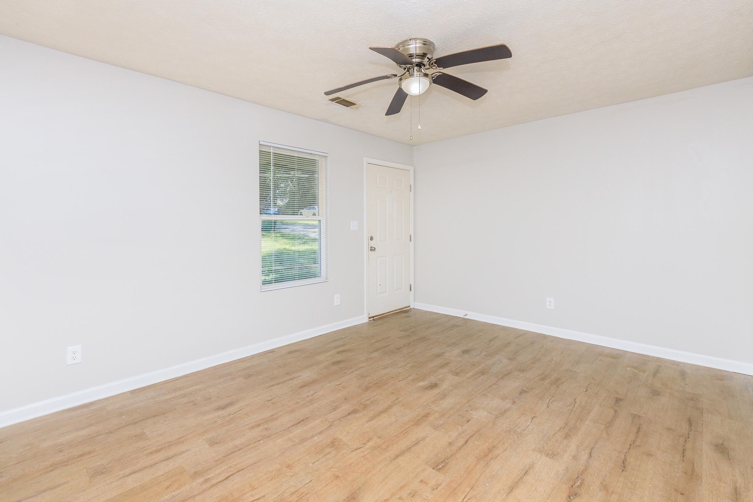 Spacious empty room with light gray walls and a ceiling fan, featuring a window and a door leading outside. The floor is light wood, creating a bright and airy atmosphere.