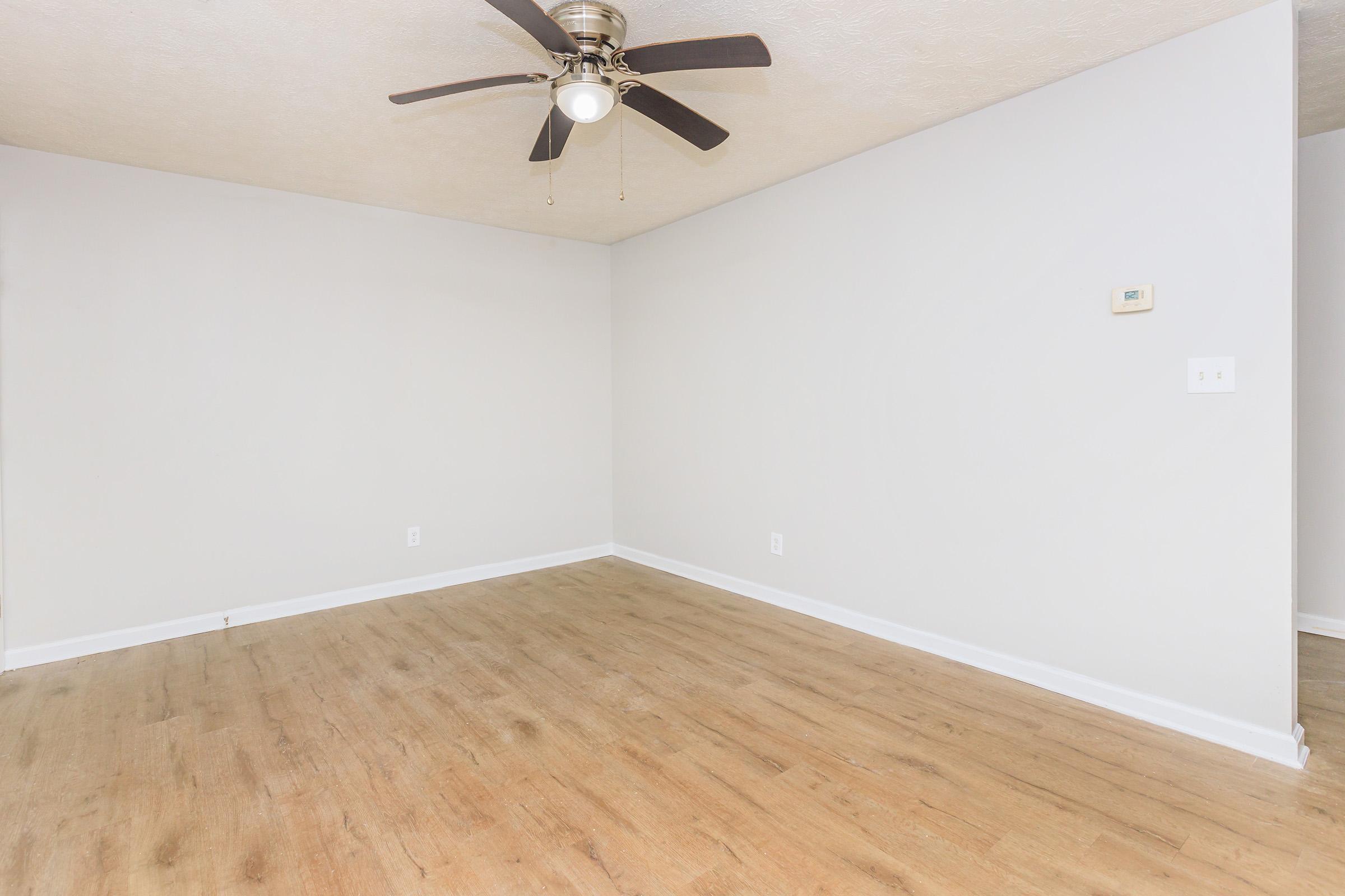 Empty room with light-colored walls and a wooden floor. A ceiling fan with four blades is mounted in the center of the ceiling. There is a small thermostat on the wall, and the room is well-lit with natural light. The space appears clean and ready for furniture.