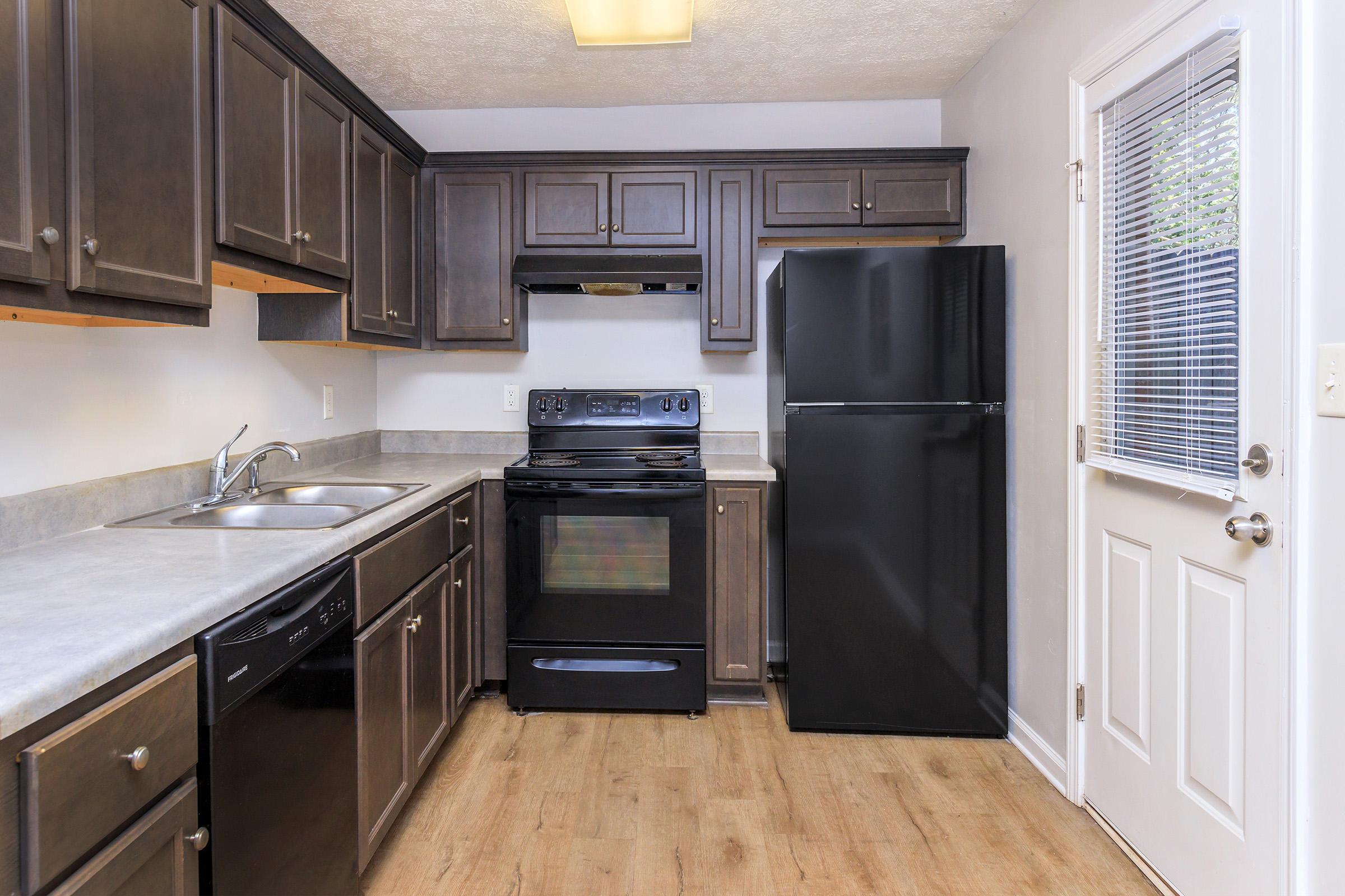 A modern kitchen featuring dark wood cabinets, a black refrigerator, and a black stove. The countertop is a light gray, and there's a stainless steel sink. A door leads outside, and a window allows natural light to enter the space. The flooring appears to be a light wood laminate.