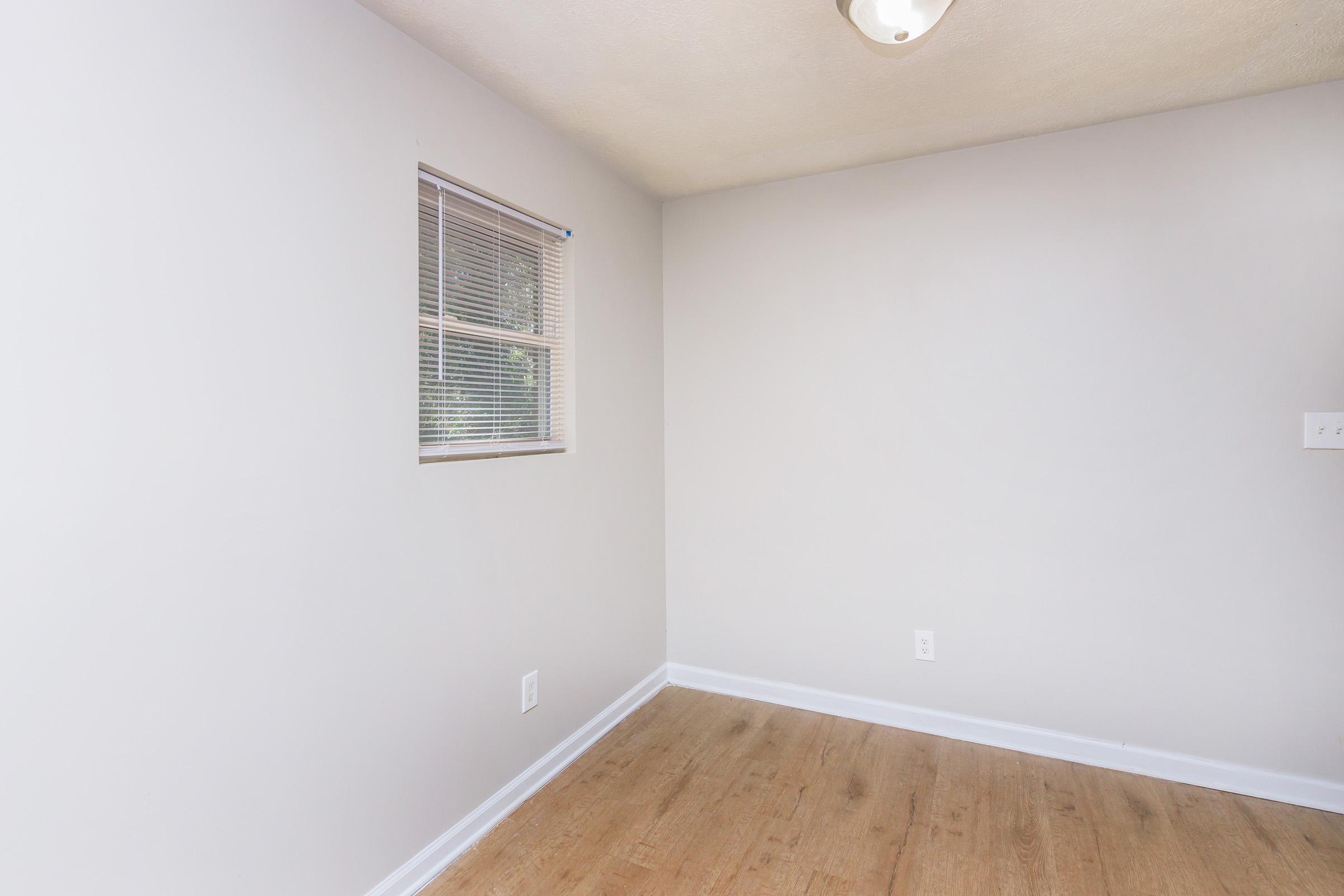 An empty room with light gray walls and a wooden floor. A small window with blinds allows natural light, and there is a ceiling fixture providing additional lighting. The space appears clean and unfurnished, ideal for various uses or decoration.