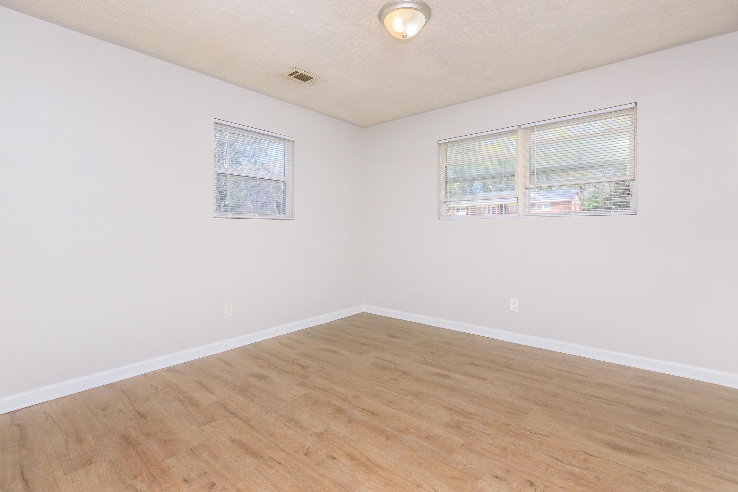 A sparsely furnished room with light gray walls, two windows allowing natural light, and a ceiling light fixture. The floor is made of light-colored wood laminate. The space appears clean and ready for potential furnishing or decoration.