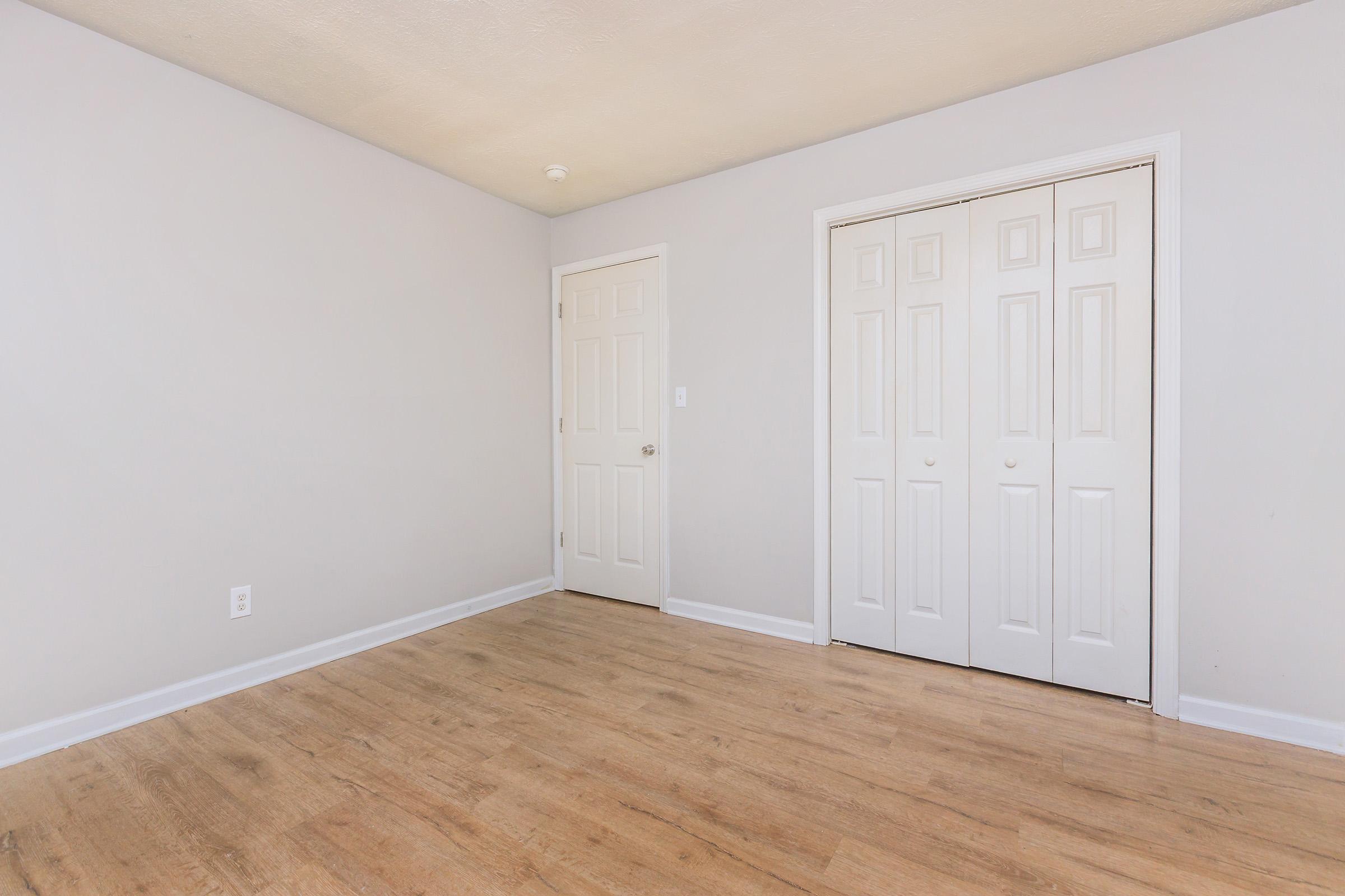 A well-lit, empty room with light gray walls and hardwood flooring. It features a closed double door closet on one side and a plain closed door on the adjacent wall, creating a minimalist and spacious aesthetic.