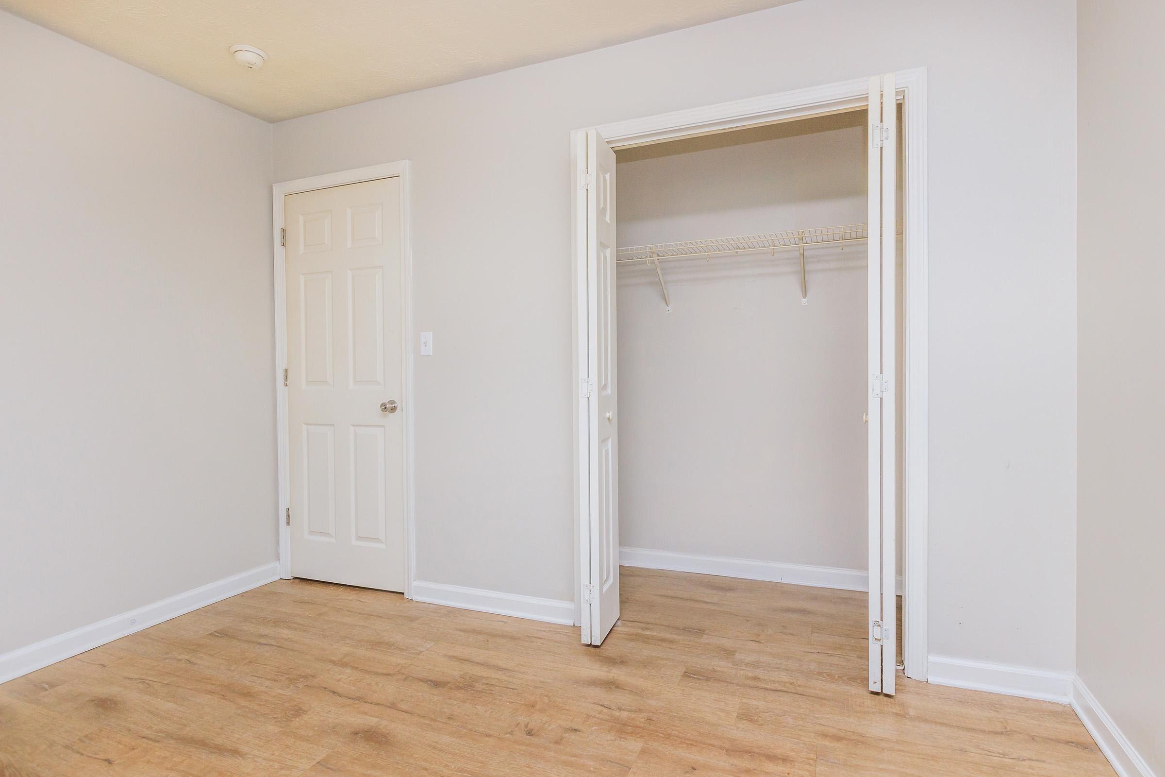 An empty room with light gray walls and a wooden floor. A door on the left leads to a closet with a wire shelf, and the closet door is open. There are no furnishings or decorations in the space, creating a clean and minimal look. Natural light brightens the room, emphasizing its simplicity.