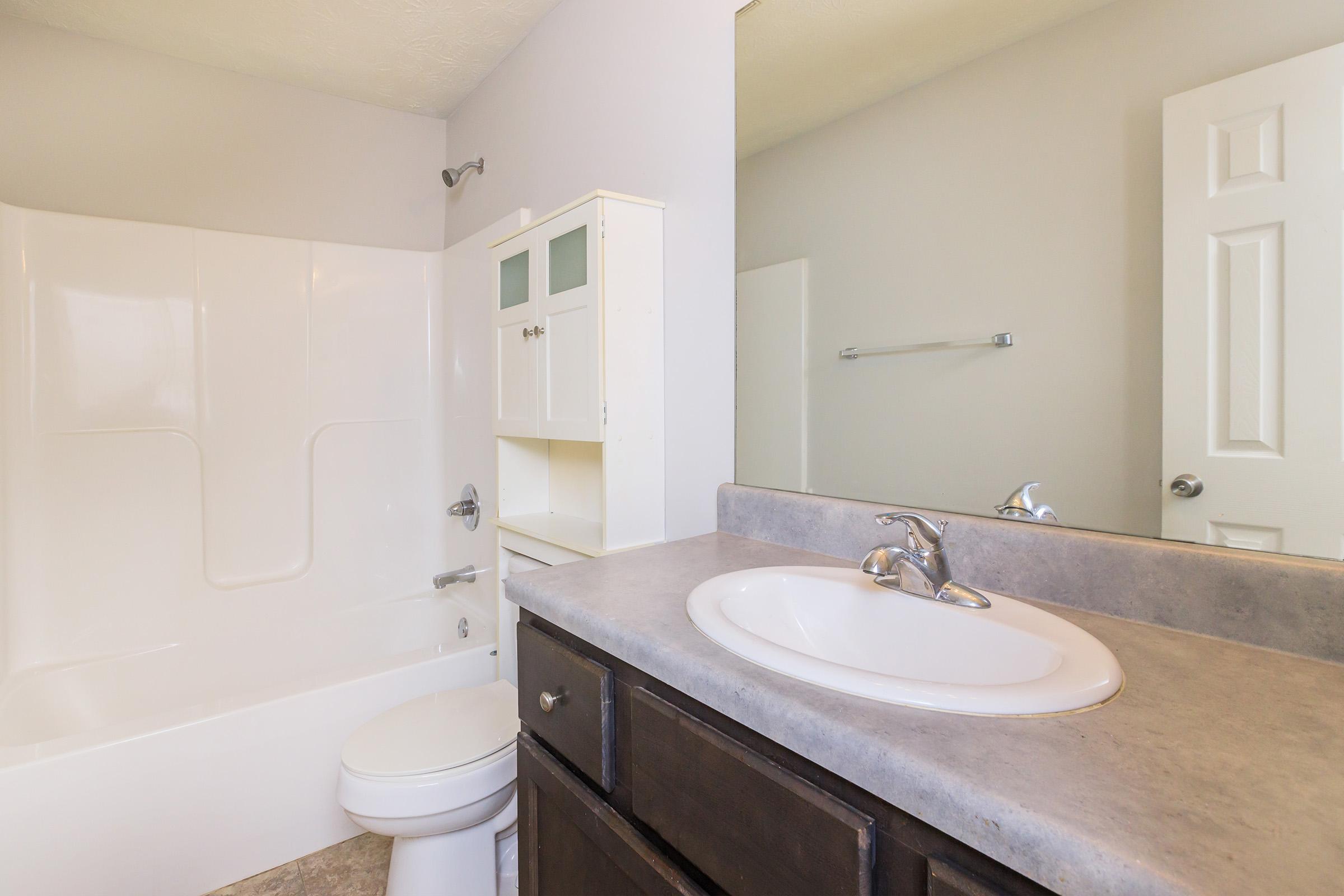 A clean, modern bathroom featuring a white bathtub with shower, a sink with a countertop, and dark wooden cabinetry. The walls are painted light gray, and there’s a mirror above the sink. A towel bar is visible, along with a light-colored door in the background.