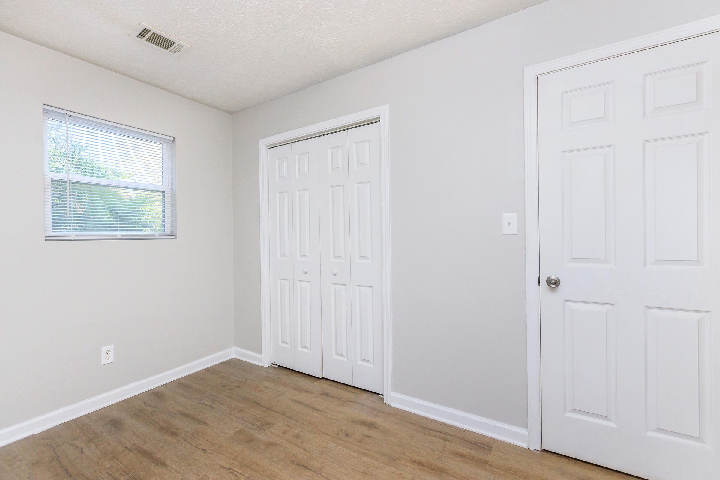 Empty room with light gray walls and a wooden floor. There is a window with blinds on the left side, and two white closet doors are located on the right wall. A simple, minimalist design with no furniture or decorations is present.