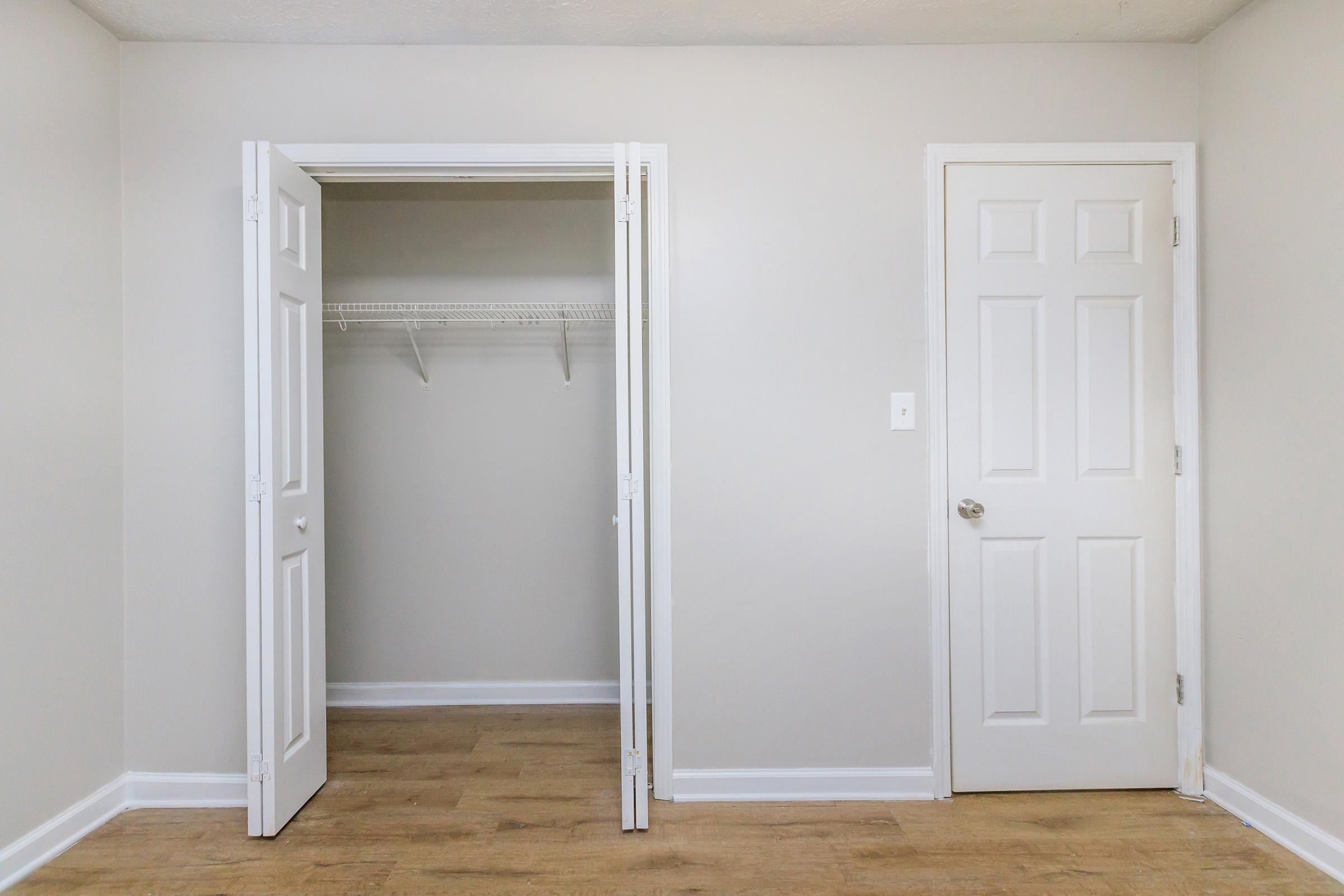 A simple, empty room with light gray walls and a wooden floor. On the left, there is an open closet with a shelf, and on the right, a closed door. The space has a clean and minimalistic design, featuring white trim and door frames.