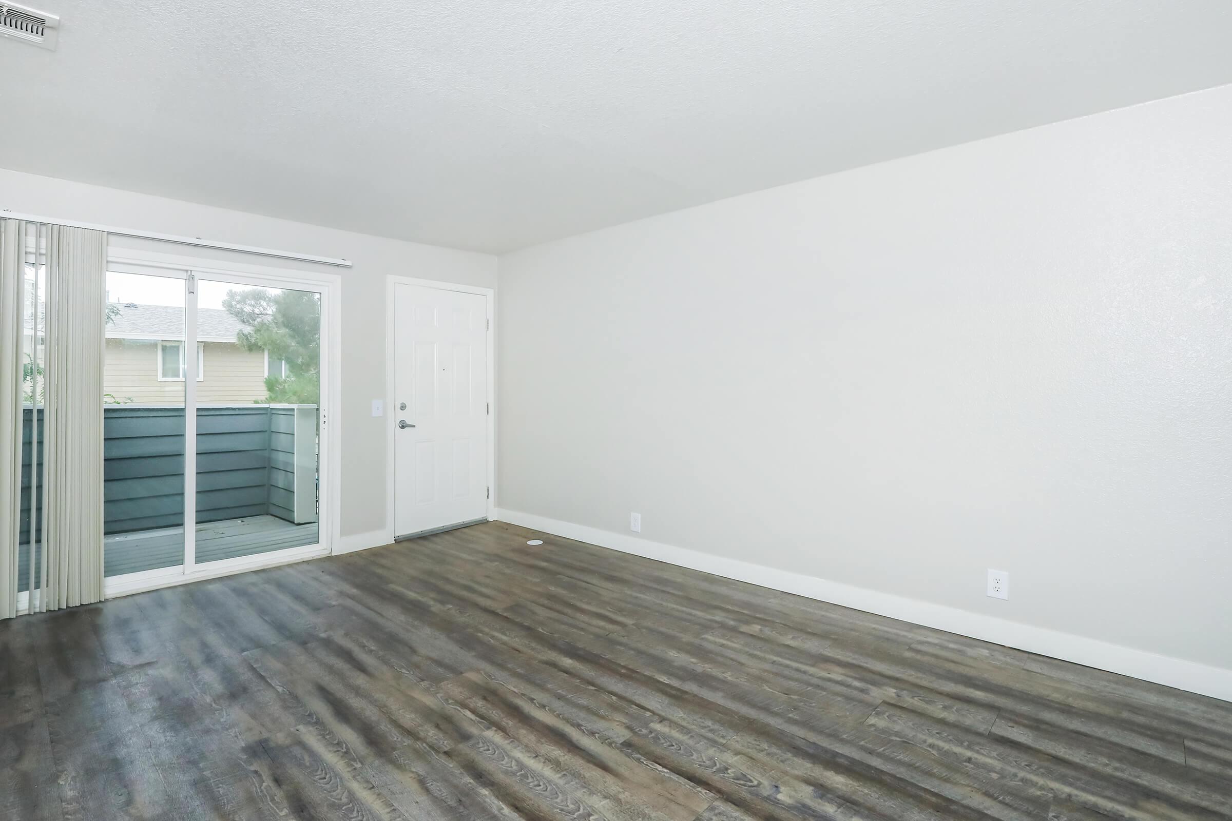 A spacious, empty room with a wooden floor and light-colored walls. There is a sliding glass door leading to a small balcony on one side. The entrance door is visible on the opposite wall, with no furniture present, creating a bright and airy atmosphere.