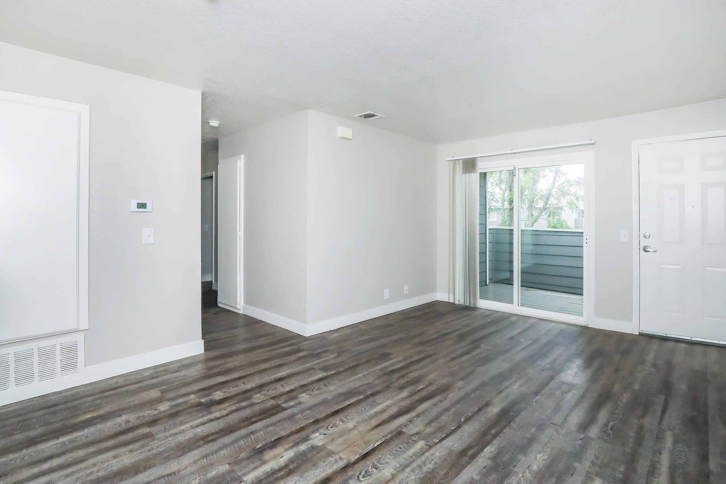 A spacious and empty living area featuring light gray walls, a sliding glass door leading to a small balcony, and brown laminate flooring. A white interior door and a small hallway are visible in the background, providing access to other rooms. Natural light is coming through the door.