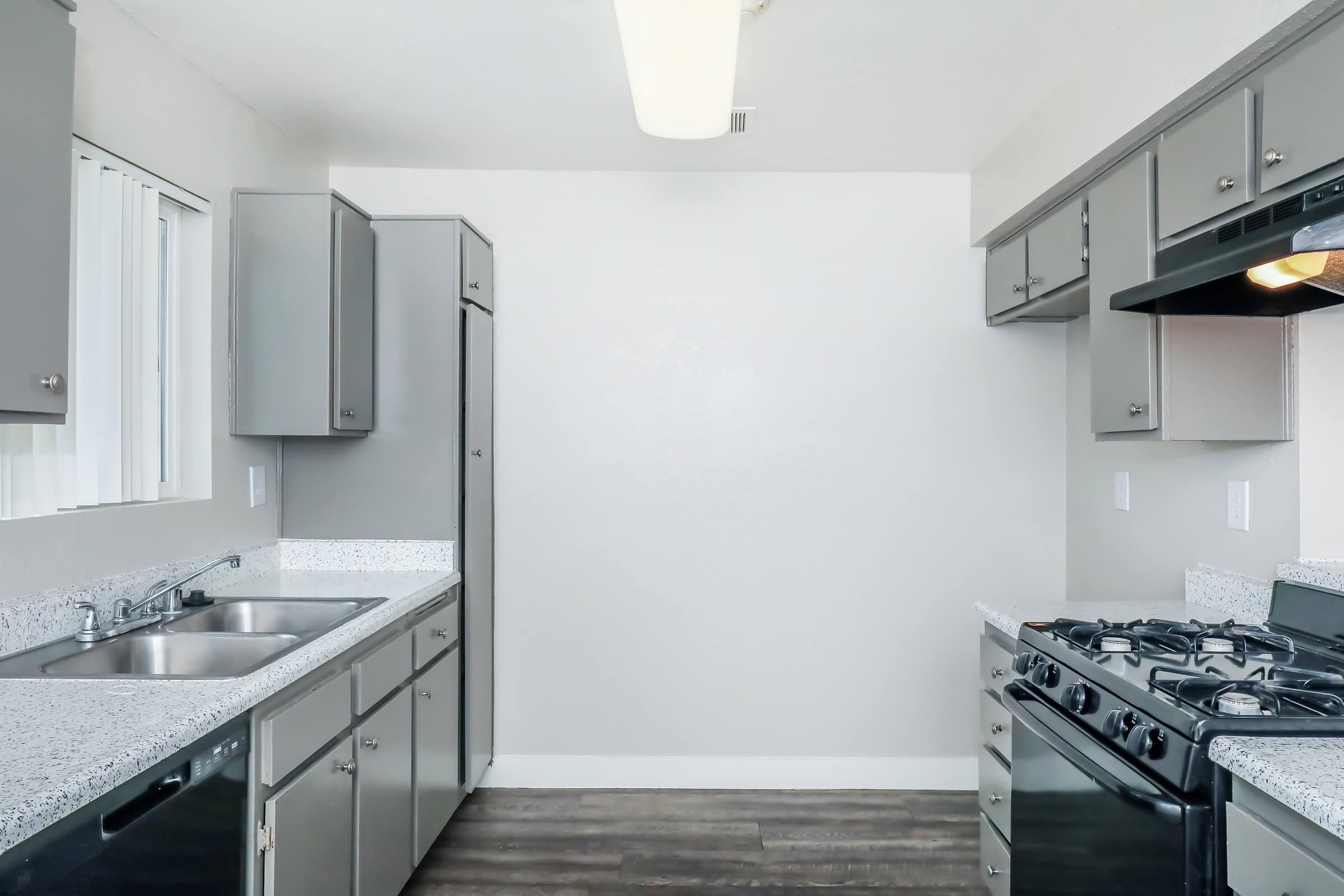 A modern kitchen featuring light gray cabinets, a white wall, a double sink, and a stove with a hood. The space includes a refrigerator, and the floor is covered with dark wood laminate. Bright lighting fixtures hang from the ceiling, enhancing the airy feel of the room.