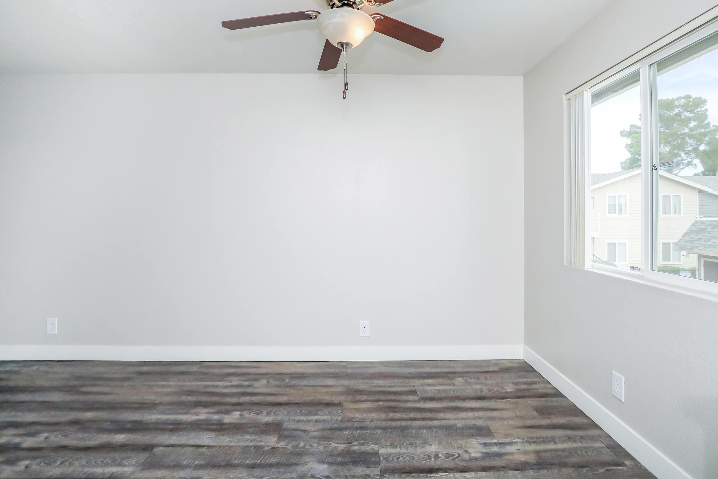 A light-colored room with a ceiling fan, featuring a large window that lets in natural light. The flooring is dark wood, and the walls are painted a neutral shade. There are no furnishings or decorations in the space, giving it a clean and minimalist appearance.