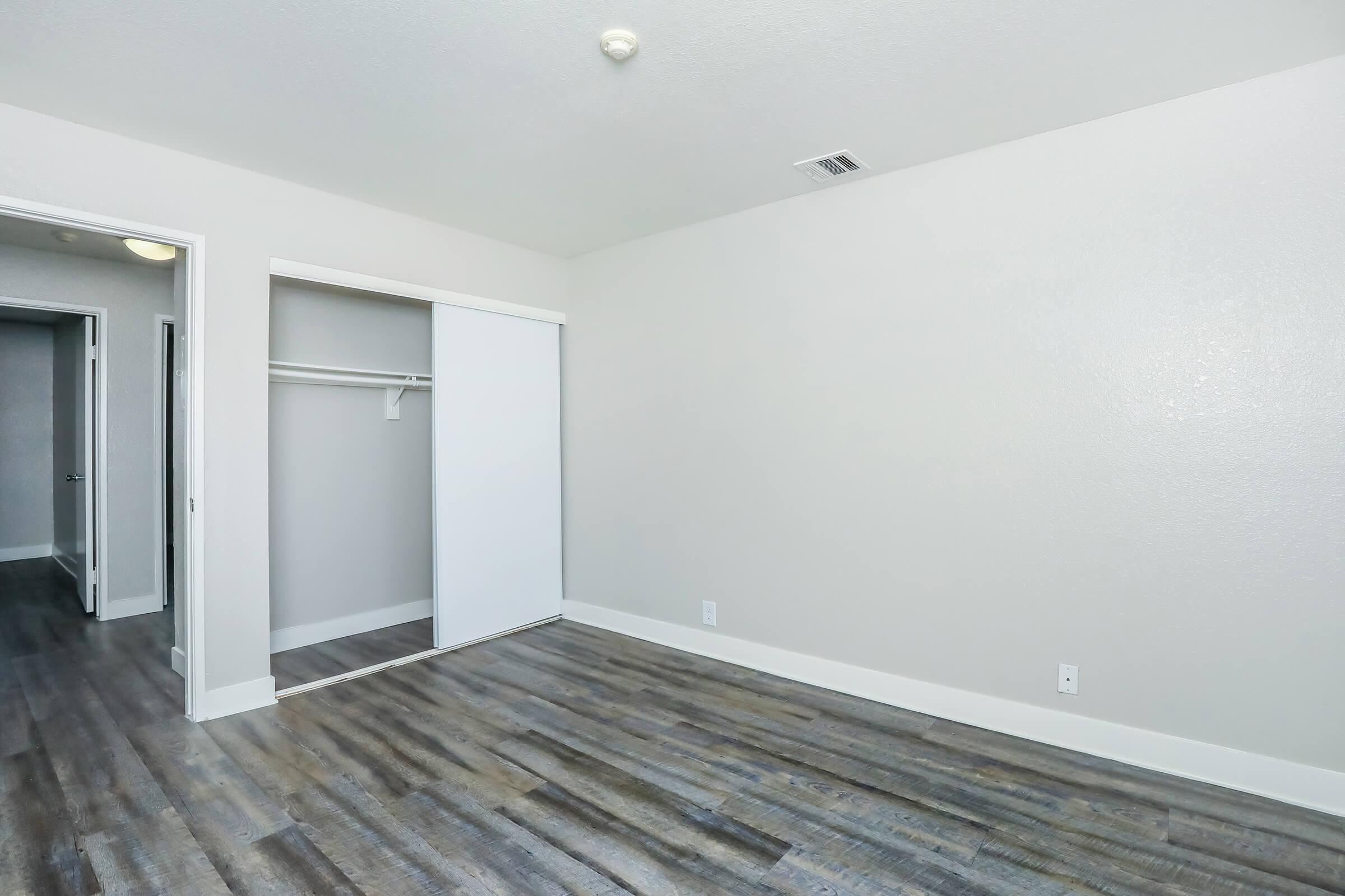 A vacant room featuring light gray walls, a wooden floor, and a sliding closet door. The space is bright and airy, with natural light coming from an unseen window. There is an adjoining doorway leading to another room. Ideal for a bedroom or office setup.